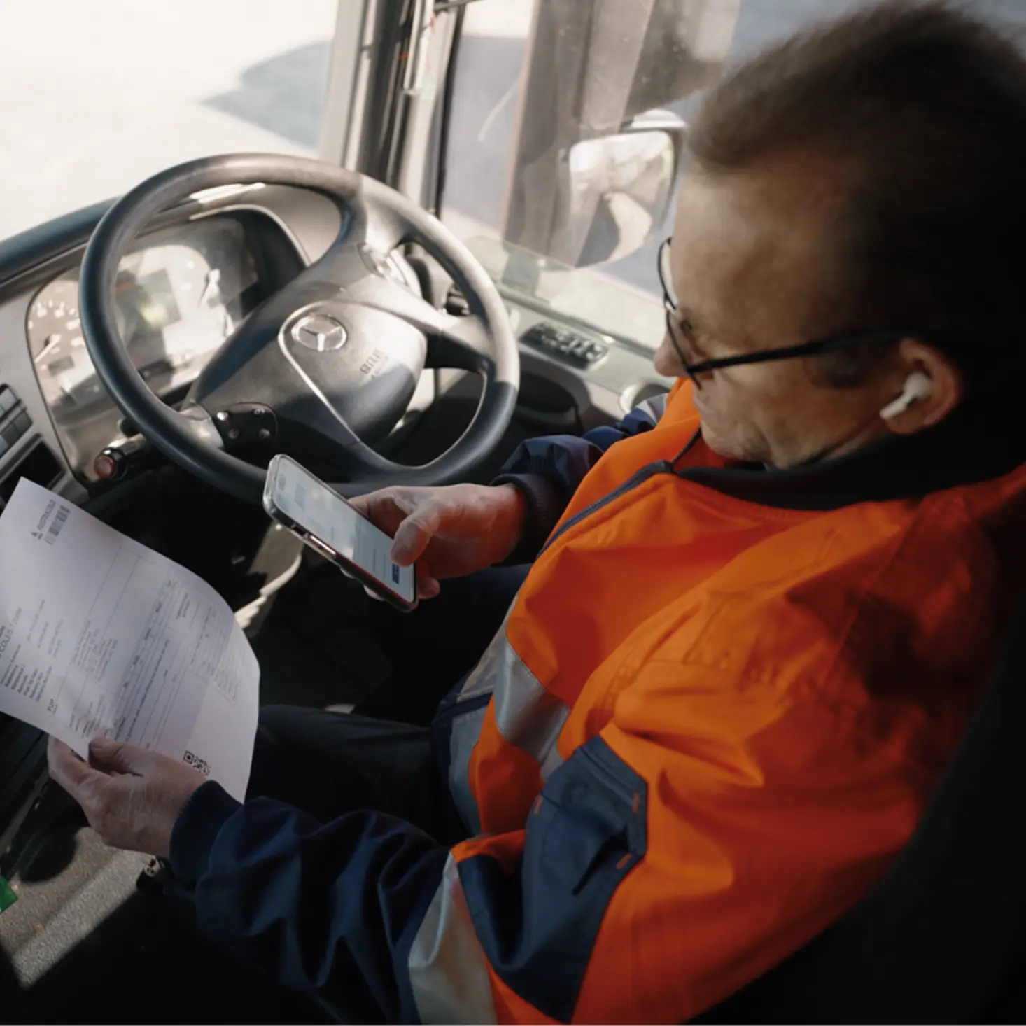 Man in an orange safety jacket sitting in a vehicle cabin, holding a document in one hand and using a smartphone with the other.