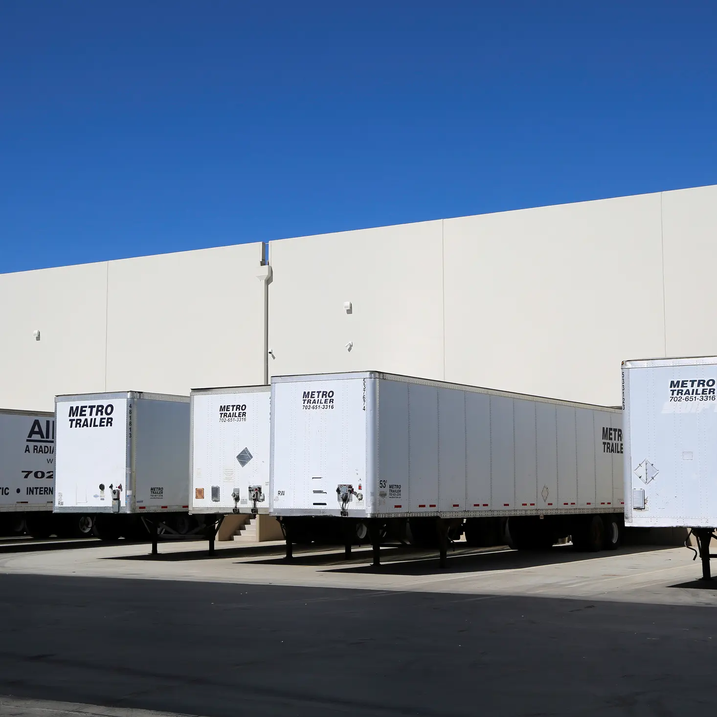 Several white Metro Trailer freight trailers parked in front of a beige warehouse under clear blue sky.