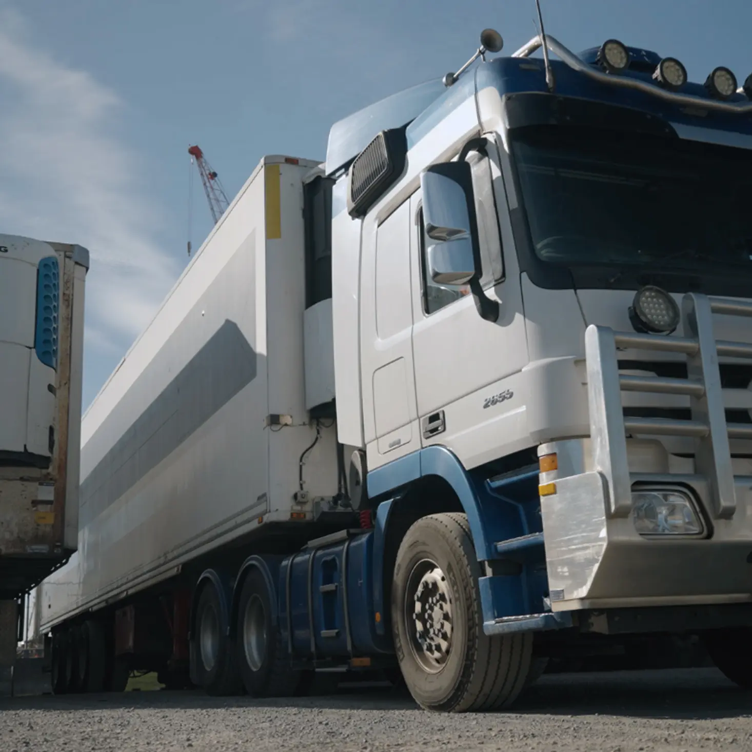 Blue and white semi-truck with a large trailer parked on a gravel surface under a partly cloudy sky.