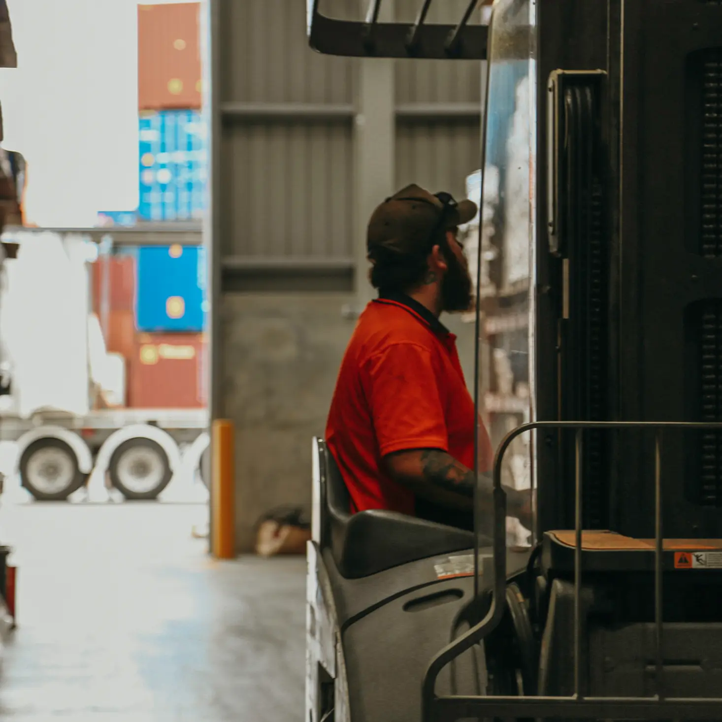 Worker in an orange shirt operating a forklift inside a warehouse with stacked shipping containers in the background.