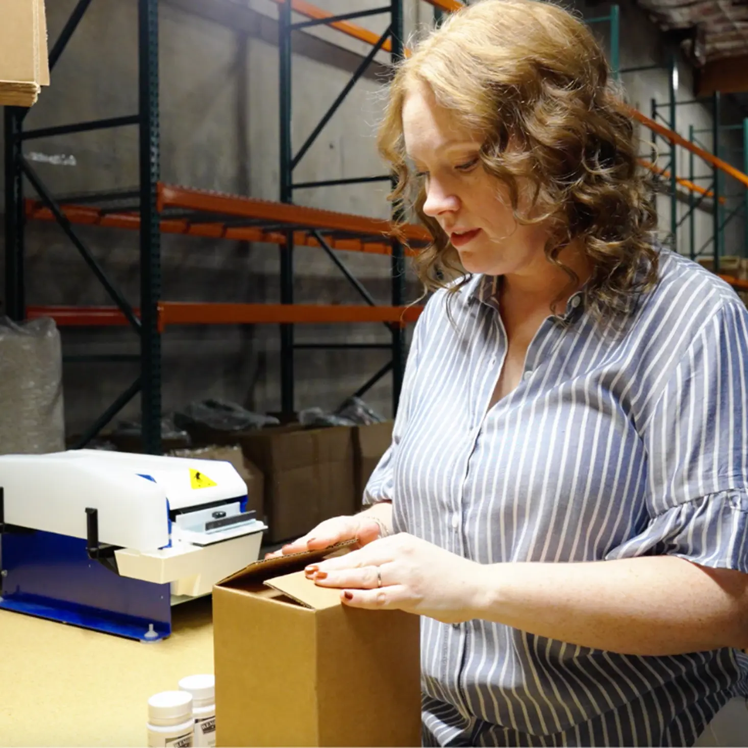 Woman with curly hair packaging a brown cardboard box in a warehouse with empty shelves and a sealing machine nearby.