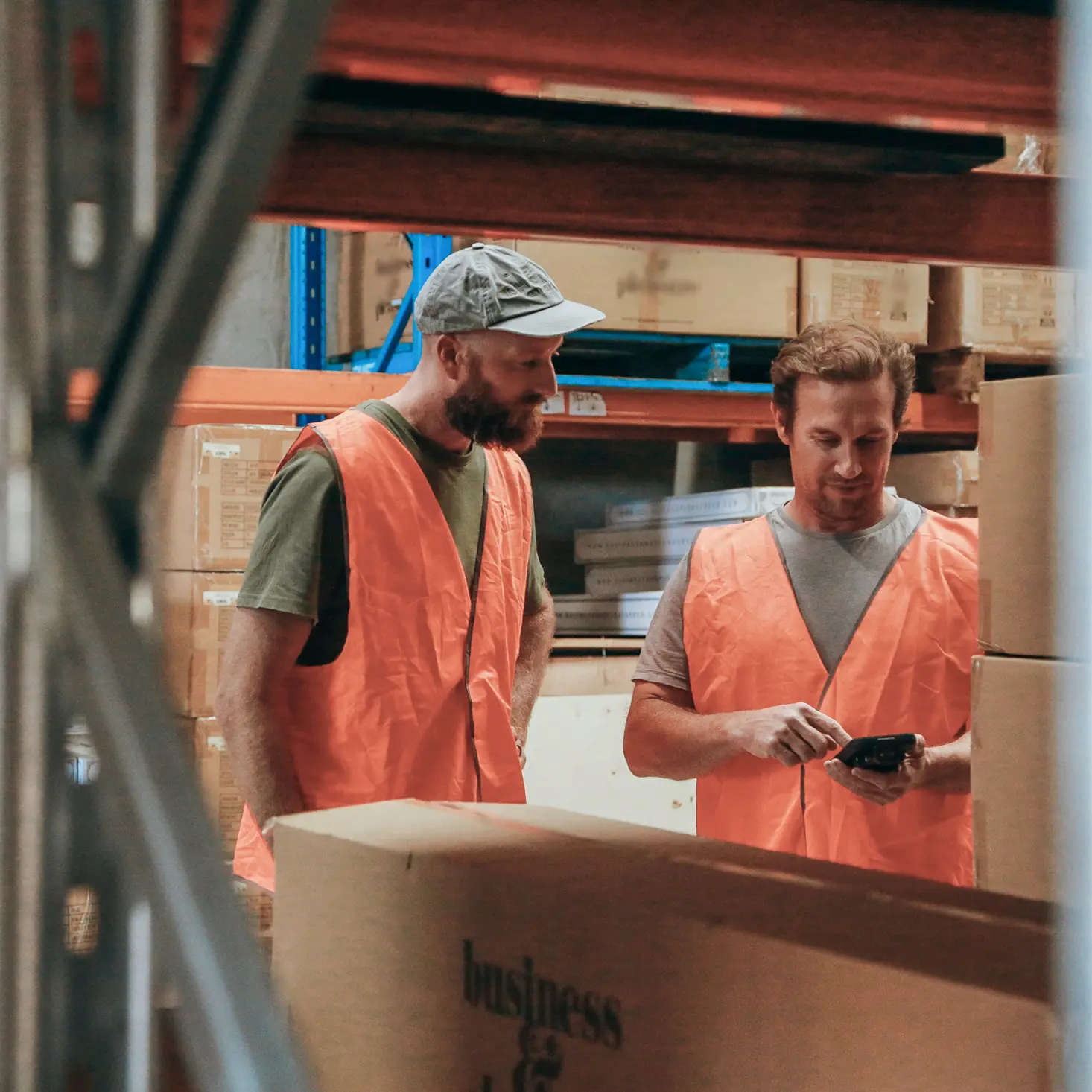 Two warehouse workers in orange safety vests inspecting a smartphone near stacked cardboard boxes.