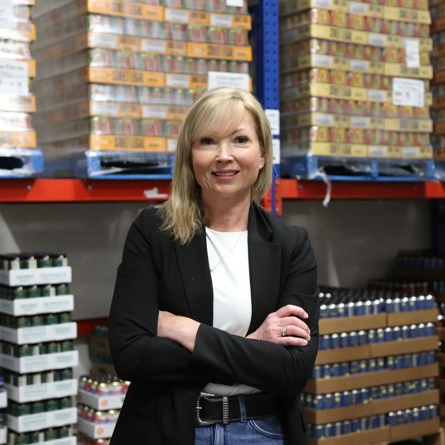 Smiling woman in a black blazer standing with arms crossed in a warehouse stocked with packaged goods.