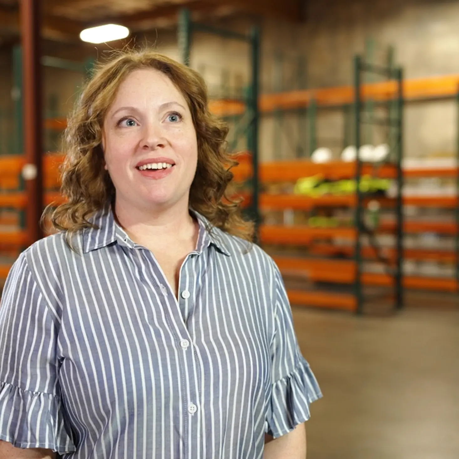 Smiling woman with curly red hair wearing a blue and white striped shirt in a warehouse with orange storage racks.