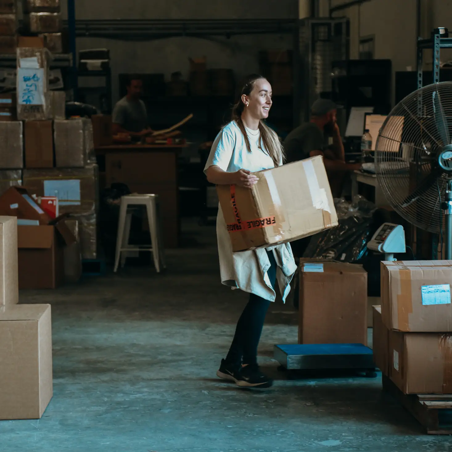 Woman carrying a cardboard box in a warehouse filled with stacked boxes and industrial equipment.