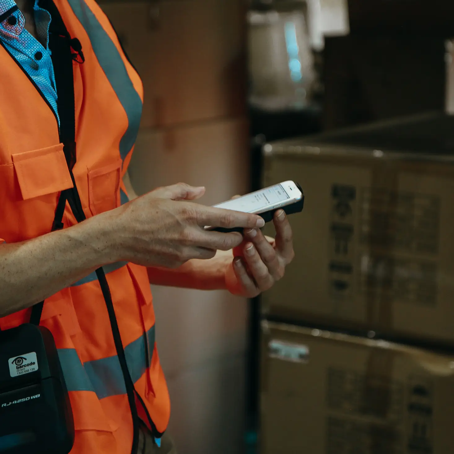 Person in orange safety vest using a handheld scanner or mobile device in a warehouse next to stacked boxes.