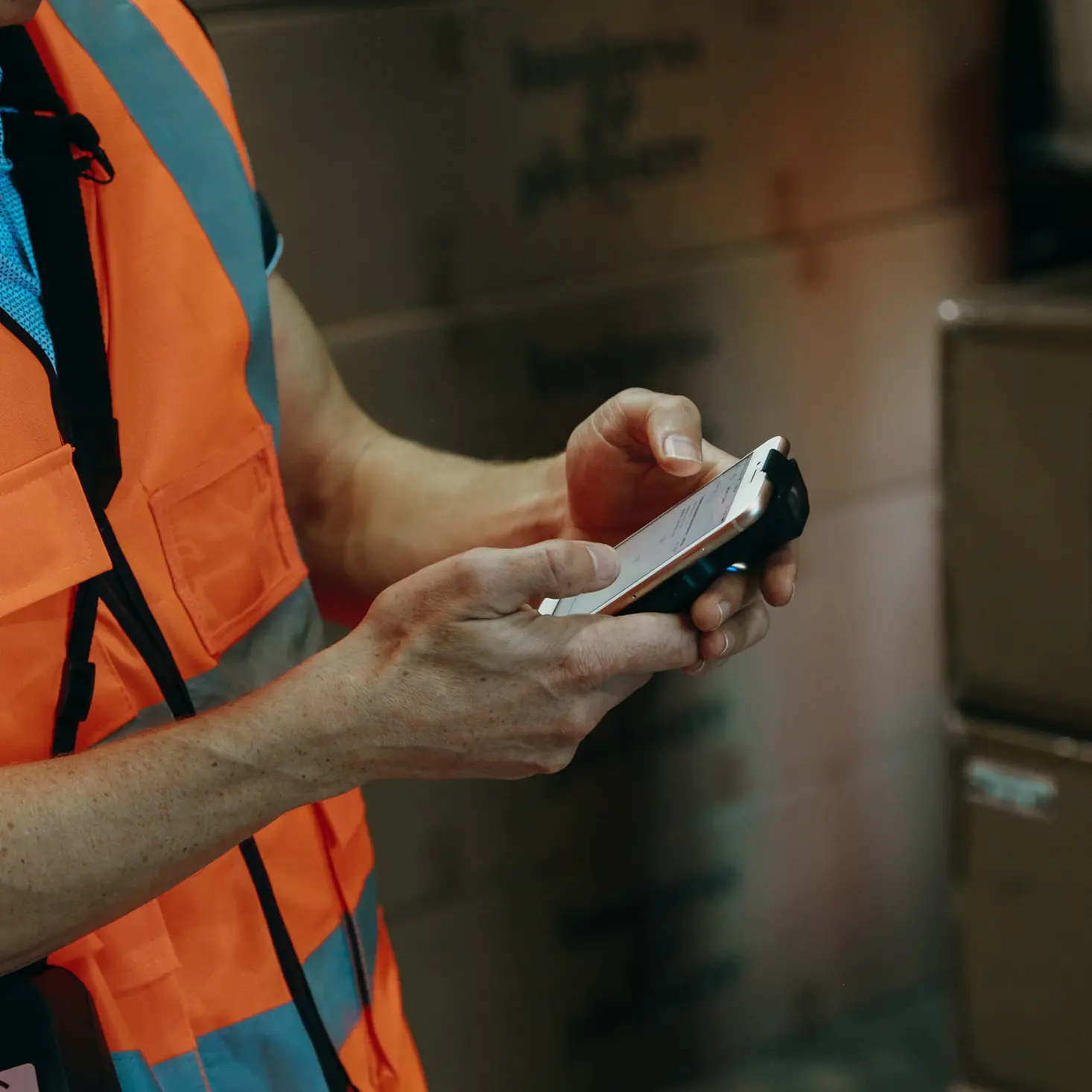 Person wearing an orange safety vest using a smartphone in a warehouse with stacked cardboard boxes in the background.