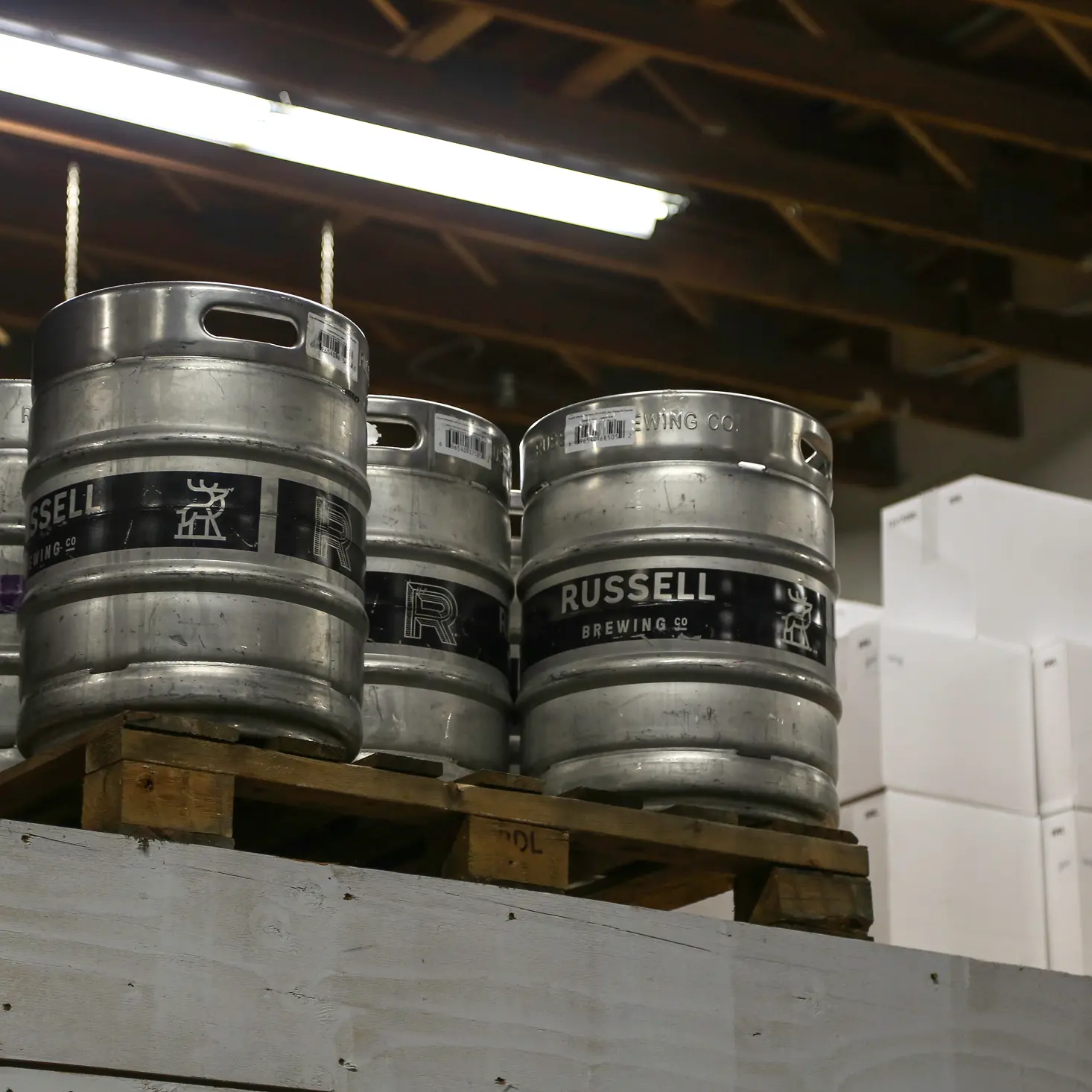 Metal kegs labeled Russell Brewing Co. stacked on a wooden pallet inside a warehouse with white boxes in the background.
