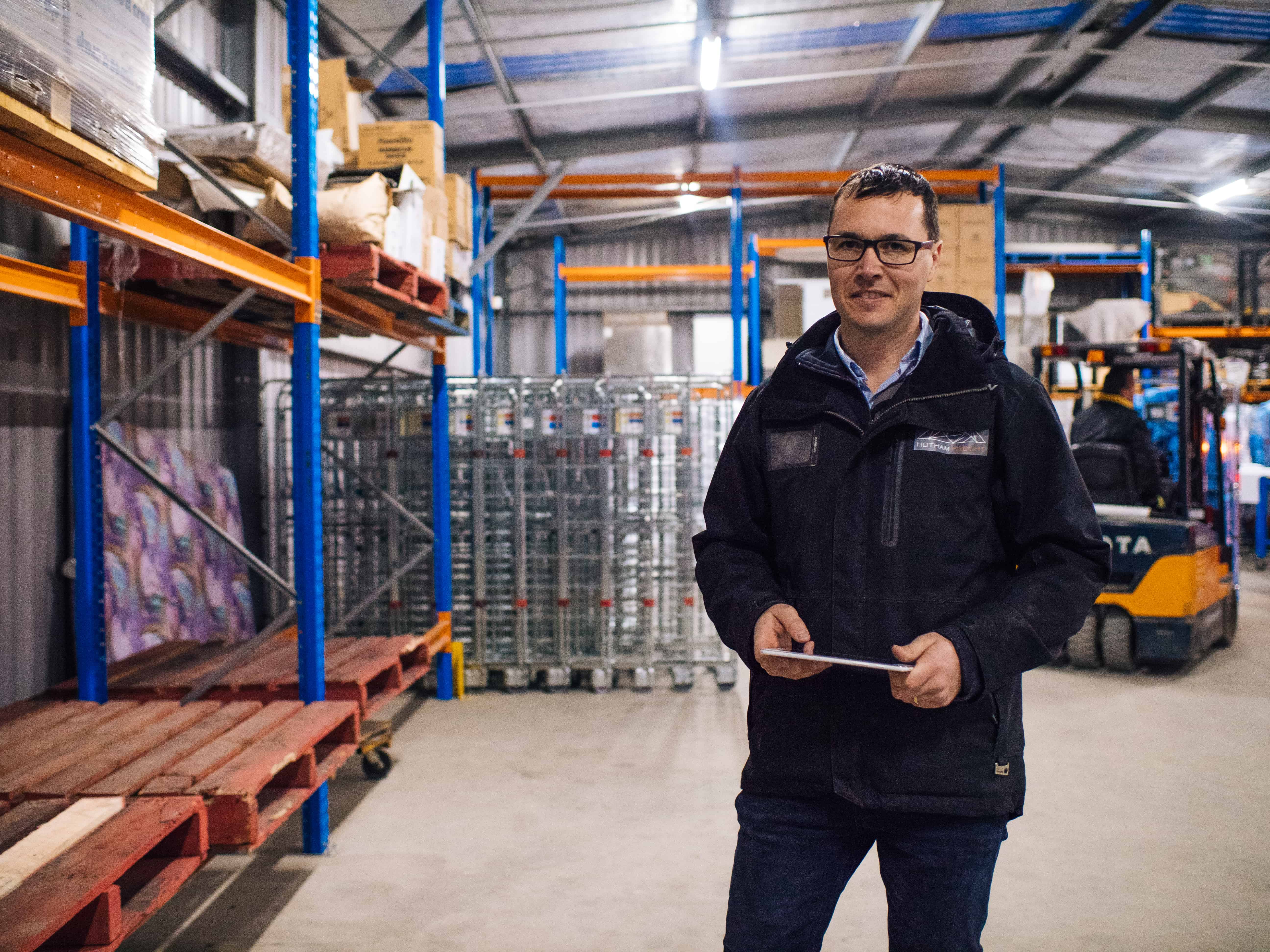 Man wearing glasses and a black jacket holding a tablet inside a warehouse with shelves and a forklift in the background.