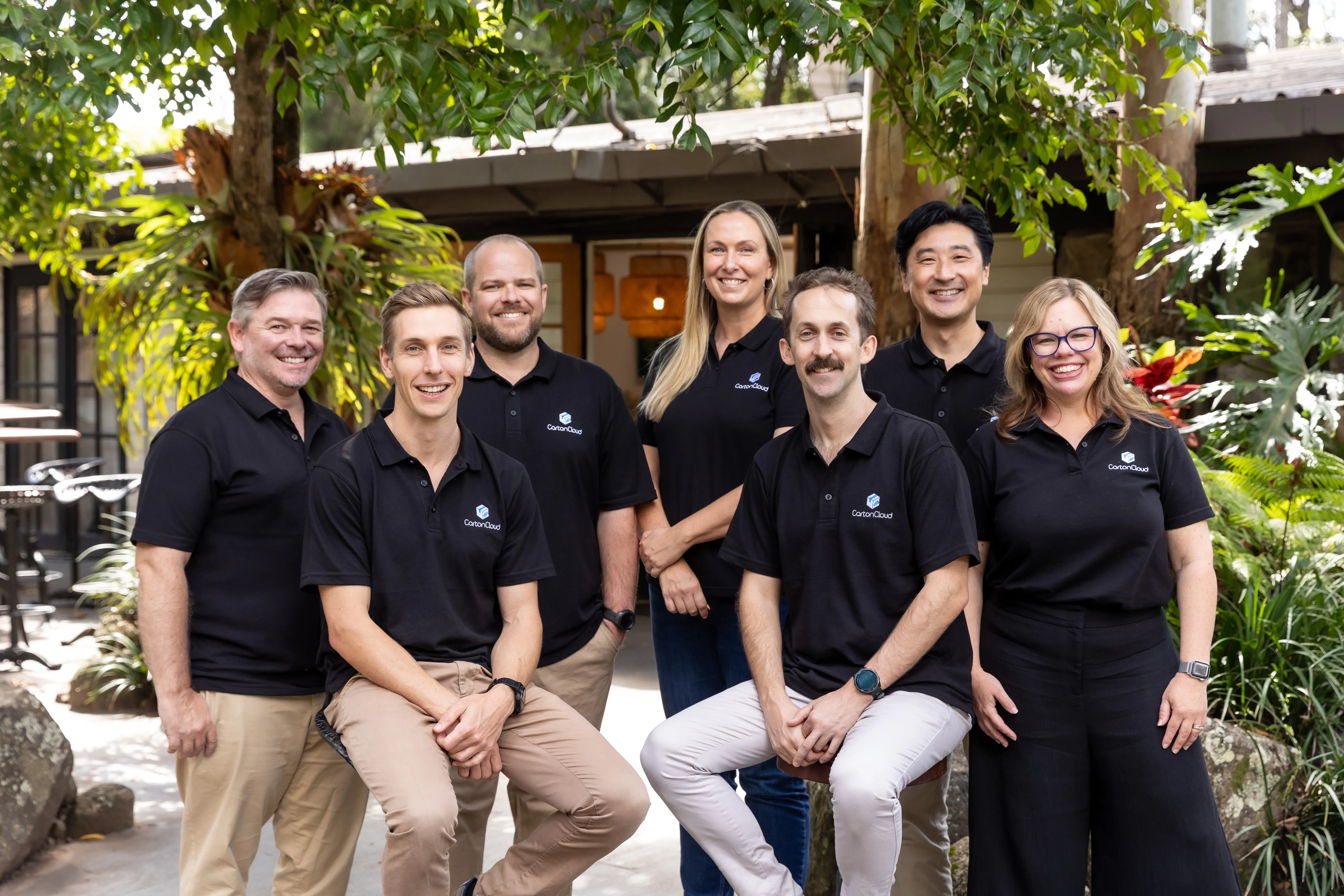 Group of seven people wearing black CartonCloud polo shirts, smiling outdoors in a garden setting.