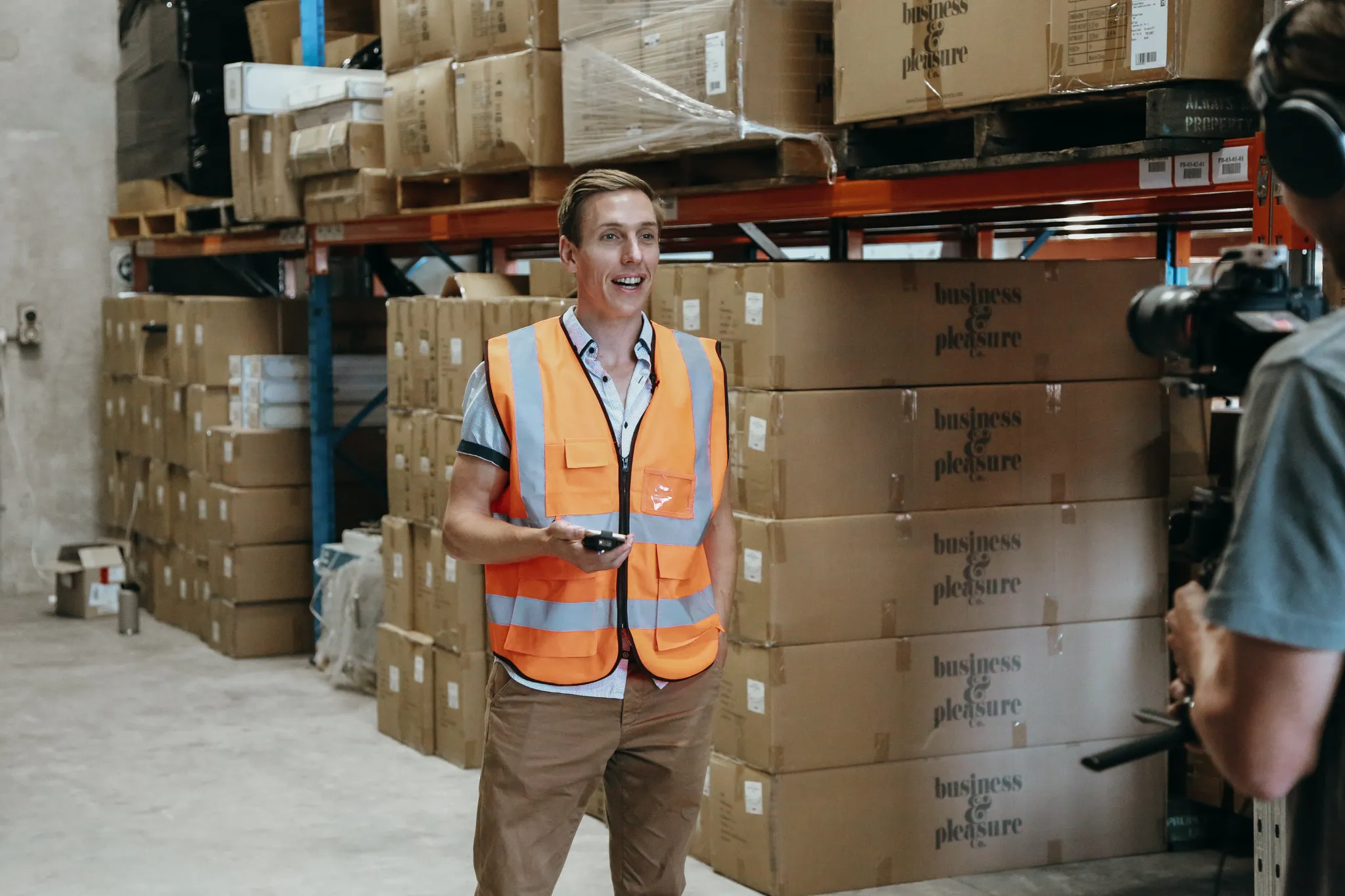Man in an orange safety vest speaking and holding a scanner in a warehouse stacked with cardboard boxes labeled Business & Pleasure Co.