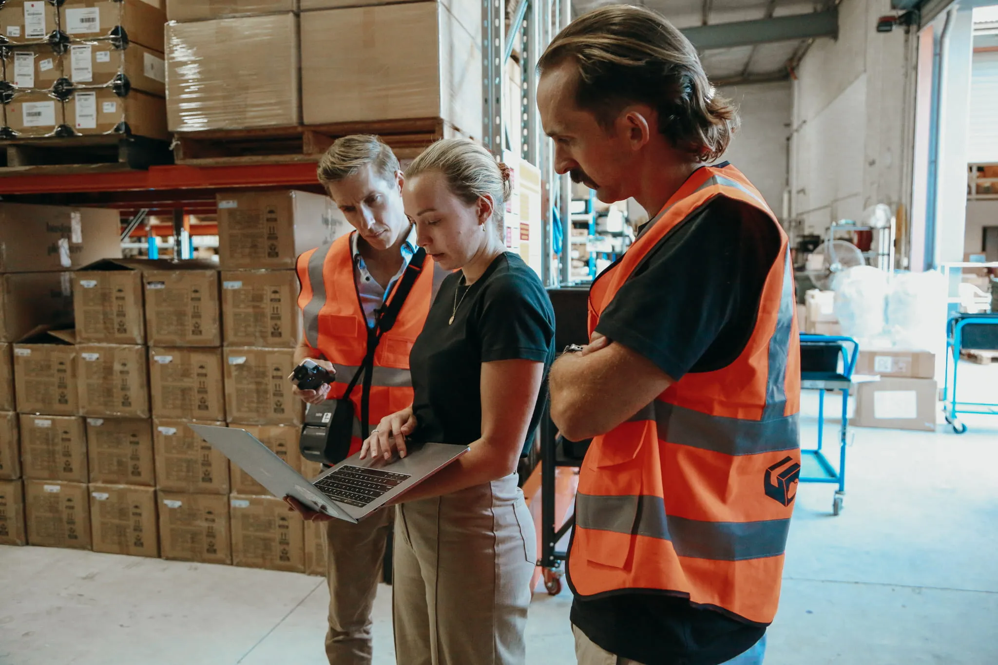 Three Cartoncloud warehouse workers, two men in orange safety vests and one woman holding a laptop, reviewing information inside a storage area with stacked boxes.