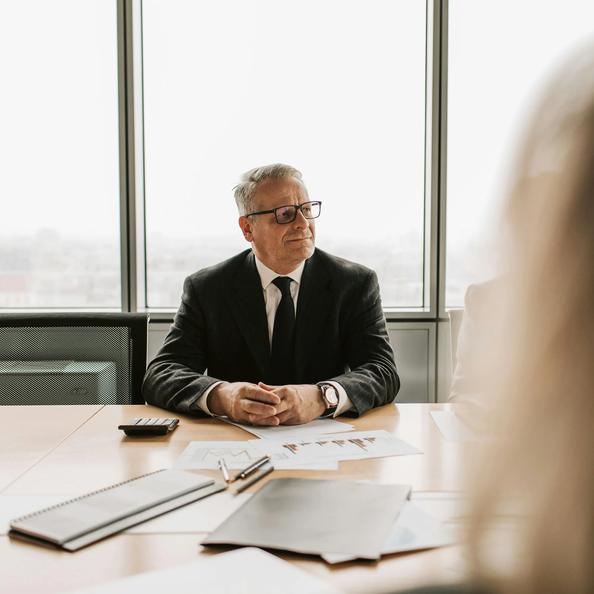 Middle-aged man in a black suit and glasses sitting at a desk with documents and a calculator, looking to the side.