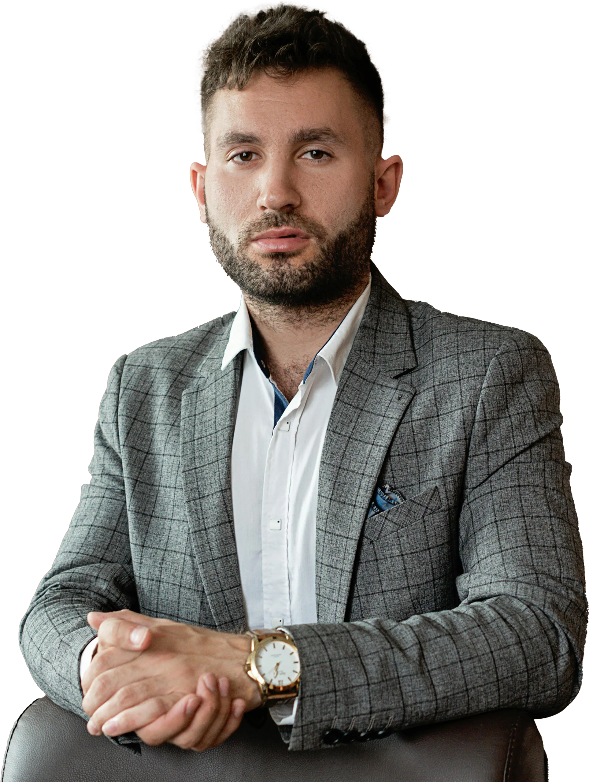 Man with short hair and beard wearing a gray checkered blazer and white shirt, resting his clasped hands on the back of a chair.