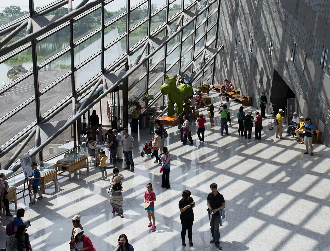 Indoor atrium with large slanted glass windows casting shadows on the floor, filled with people walking, sitting, and viewing exhibits including a green sculpture.