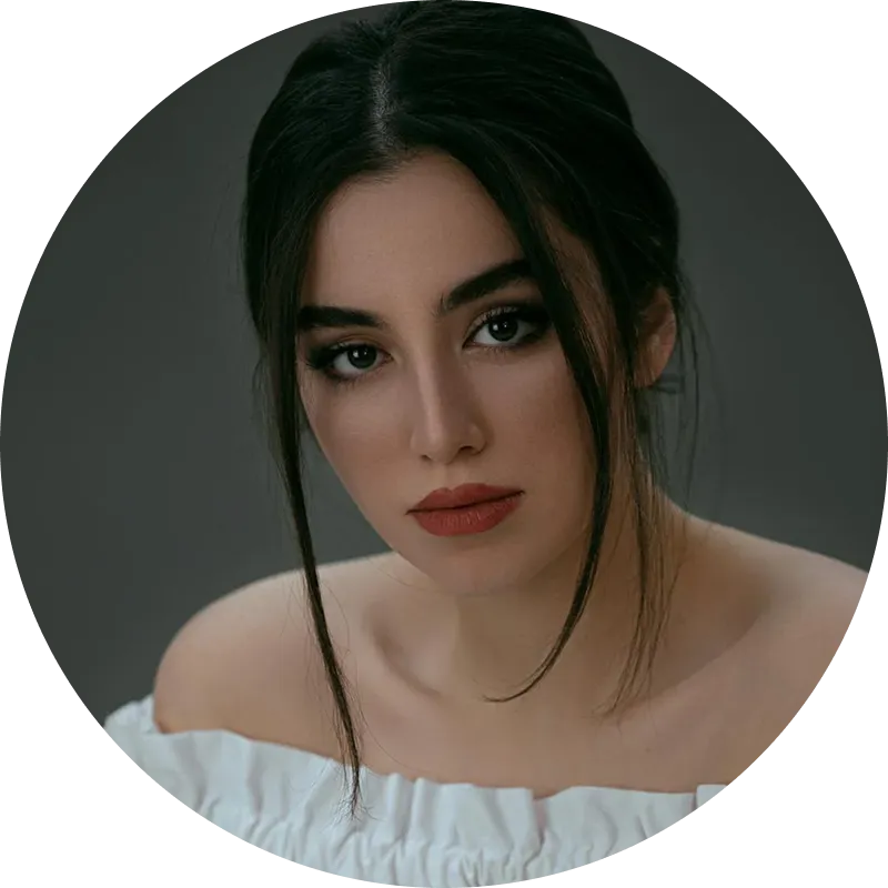 Portrait of young woman with dark hair and brown eyes wearing an off-shoulder white top against a dark background.