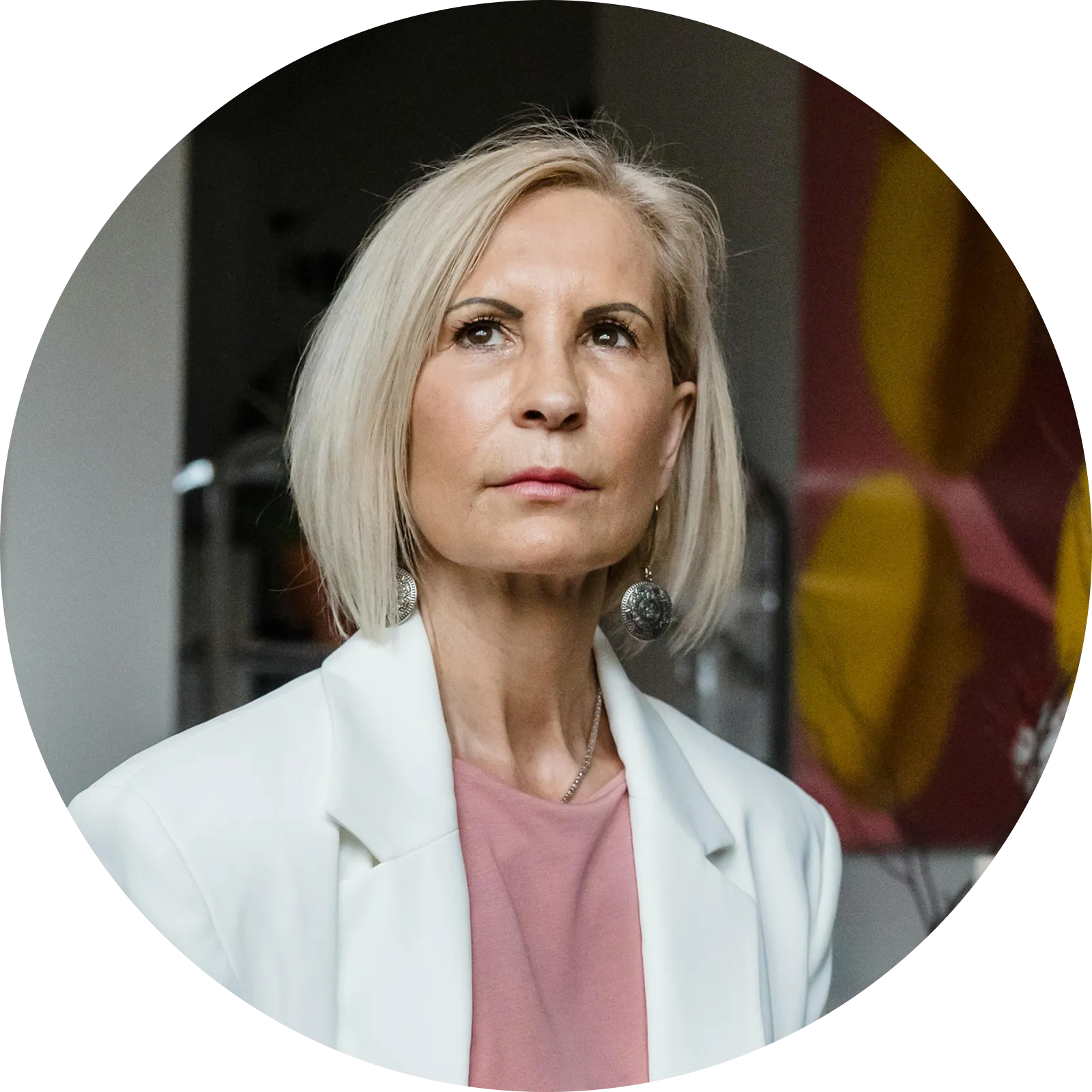 Thoughtful middle-aged woman with short blonde hair wearing a white blazer and pink top indoors.