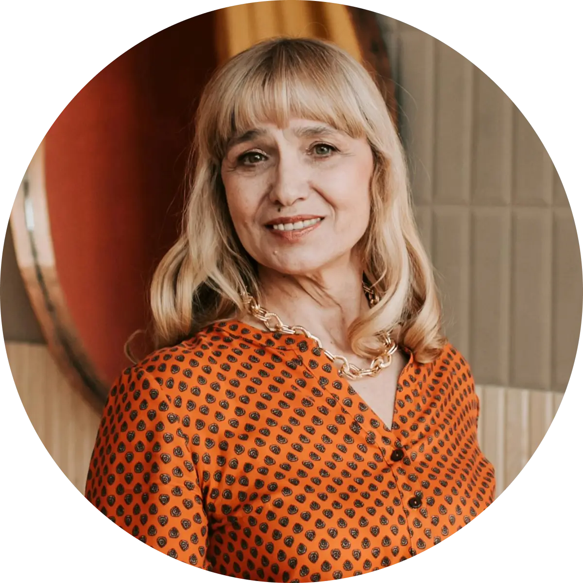 Smiling woman with blonde hair wearing an orange patterned blouse and a gold chain necklace.