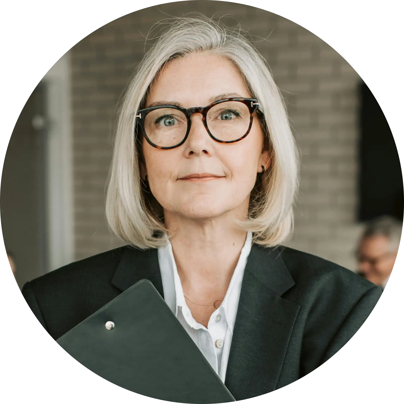 Professional woman with gray hair and glasses holding a black folder, wearing a dark blazer and white shirt.