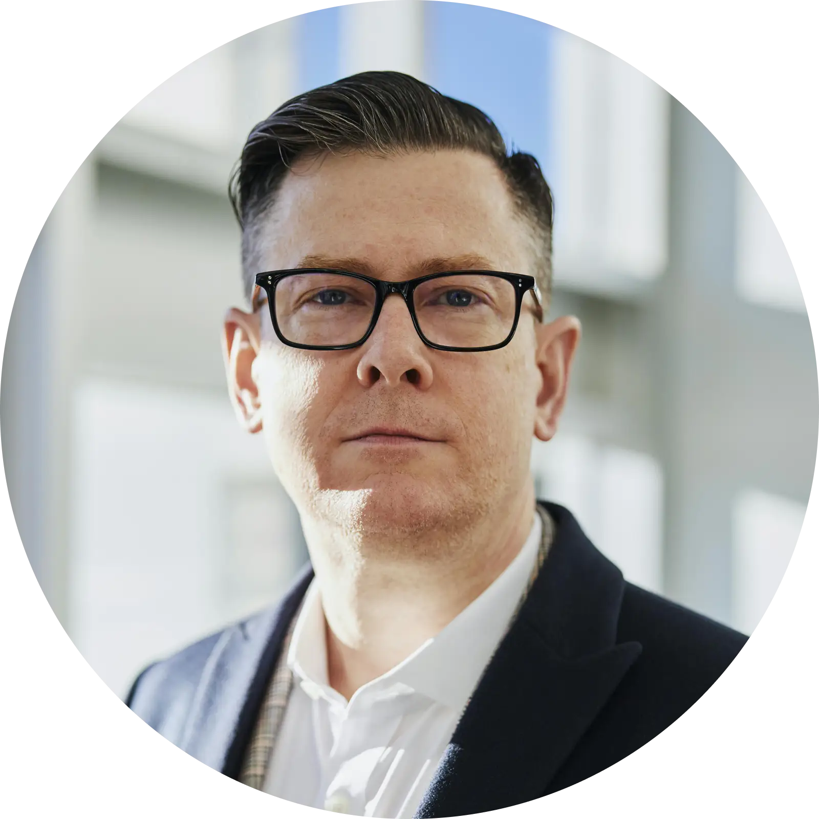 Man wearing black-rimmed glasses, white shirt, and dark blazer, looking directly at the camera with a neutral expression.