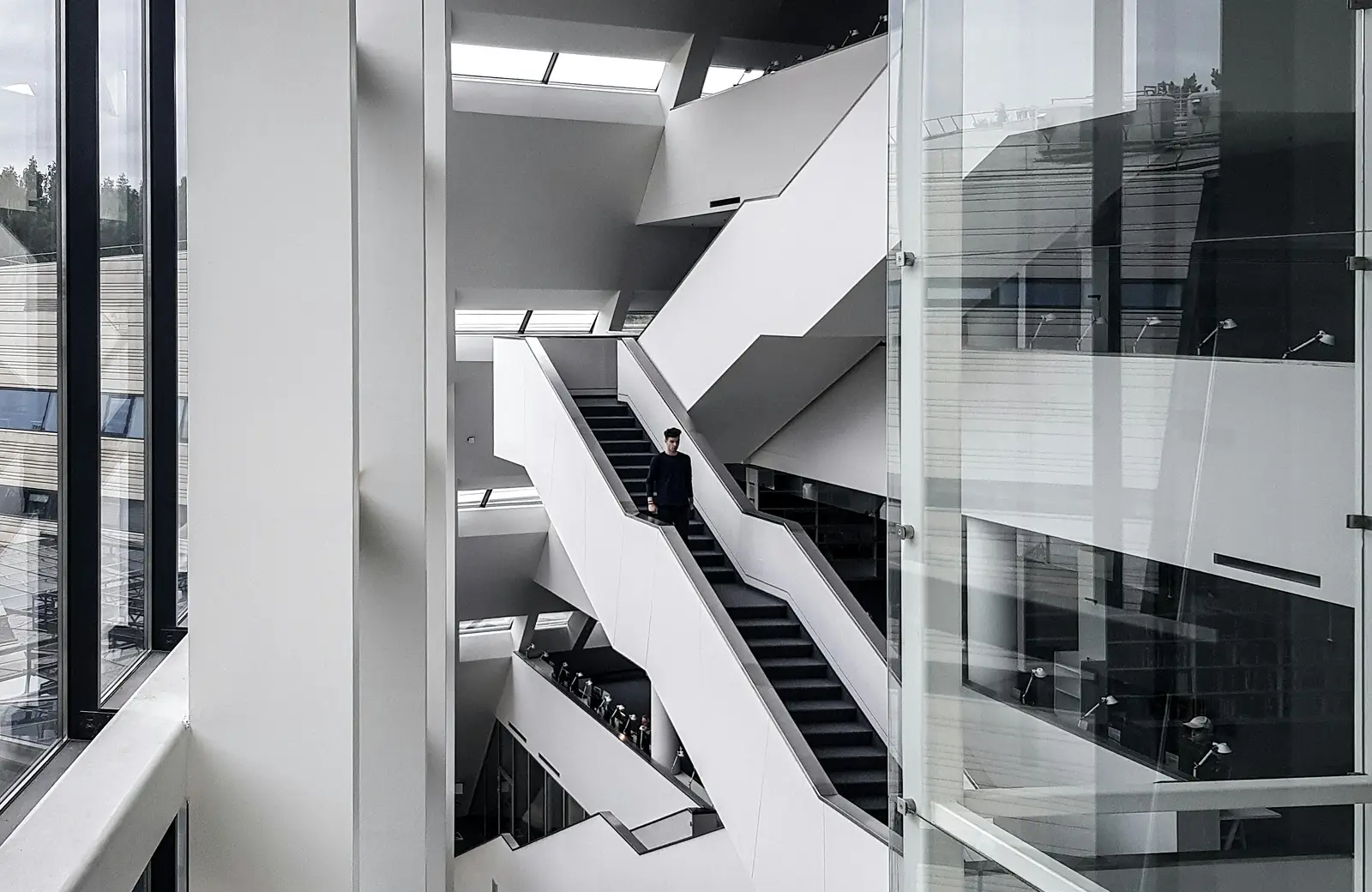A person descending a modern white staircase inside a building with large glass windows and multiple levels.