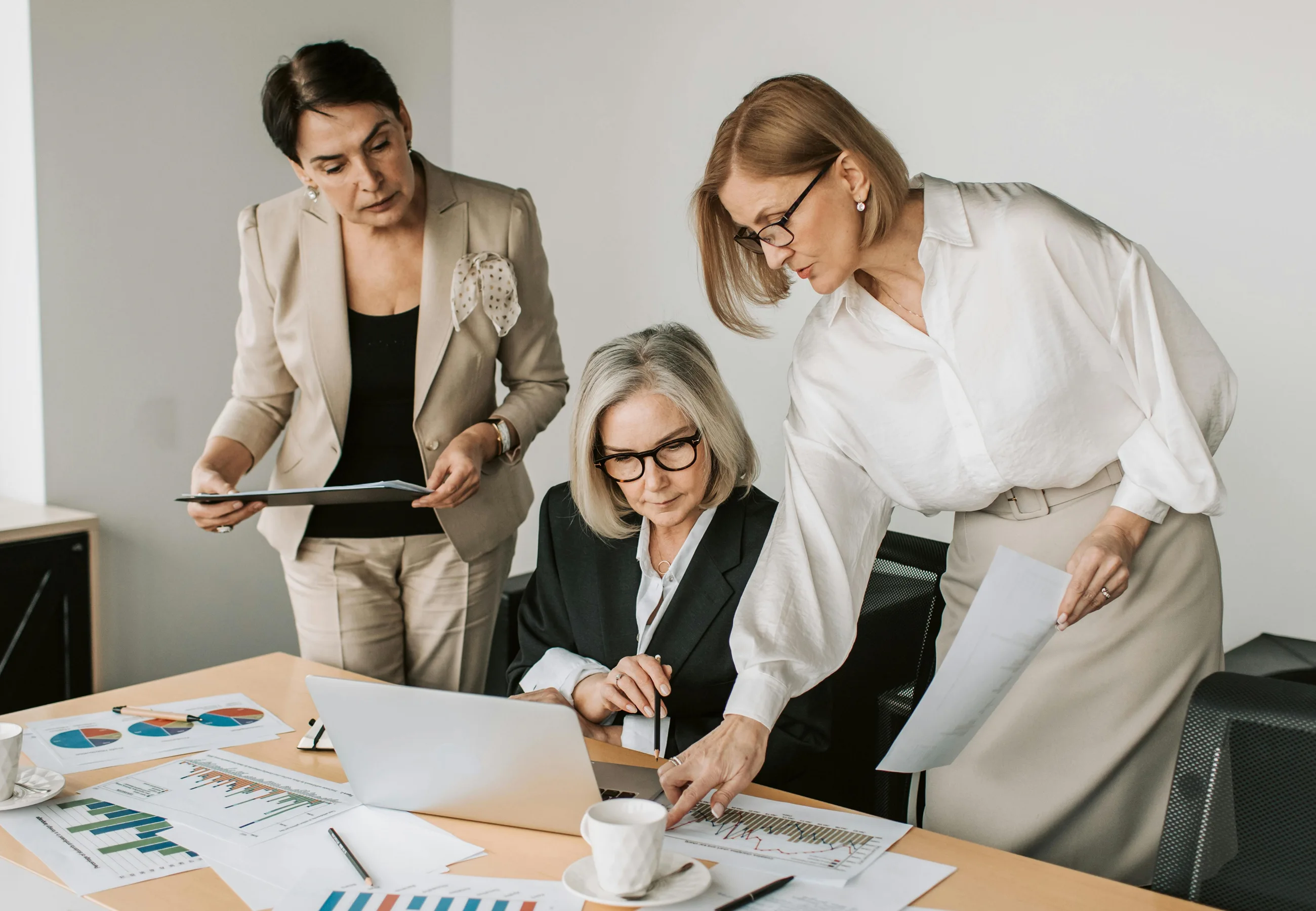 Three professional women in an office reviewing charts and documents around a laptop.