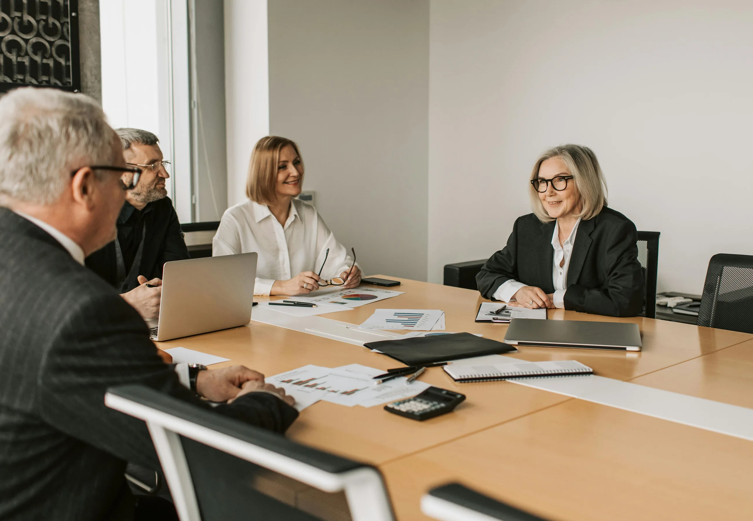 Four business professionals in a meeting room sitting around a table with laptops, documents, and charts, engaging in discussion.