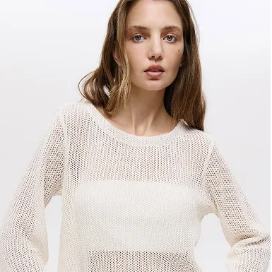 Жінка з прямим каштановим волоссям у світлому прозорому в'язаному светрі на світлому фоні.