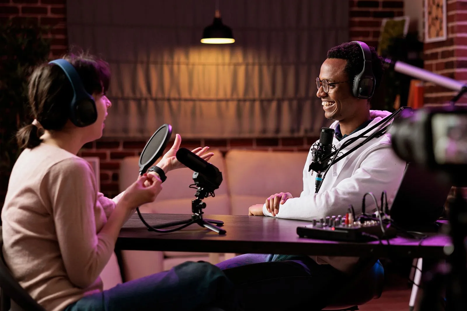 Un hombre y una mujer están haciendo un podcast, están sonriendo y pasandola bien, Hay una cámara que graba y la luz es suave y acogedora.