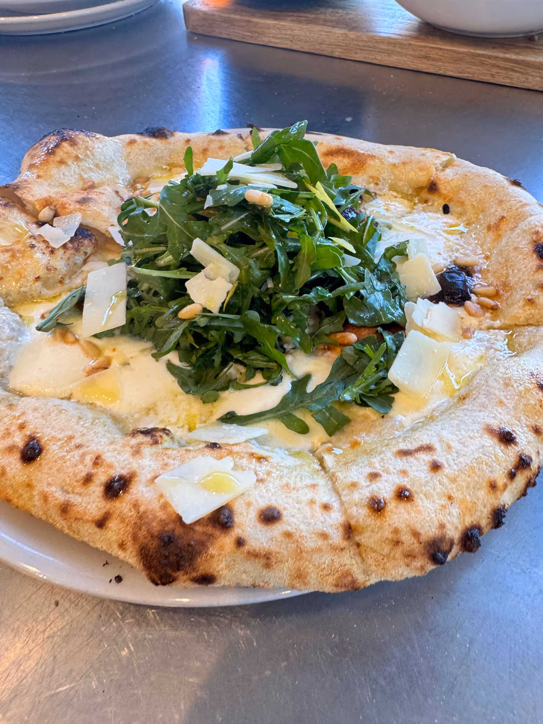 A rustic pizza topped with fresh arugula, shaved Parmesan, pine nuts, and a drizzle of olive oil sits on a white plate.