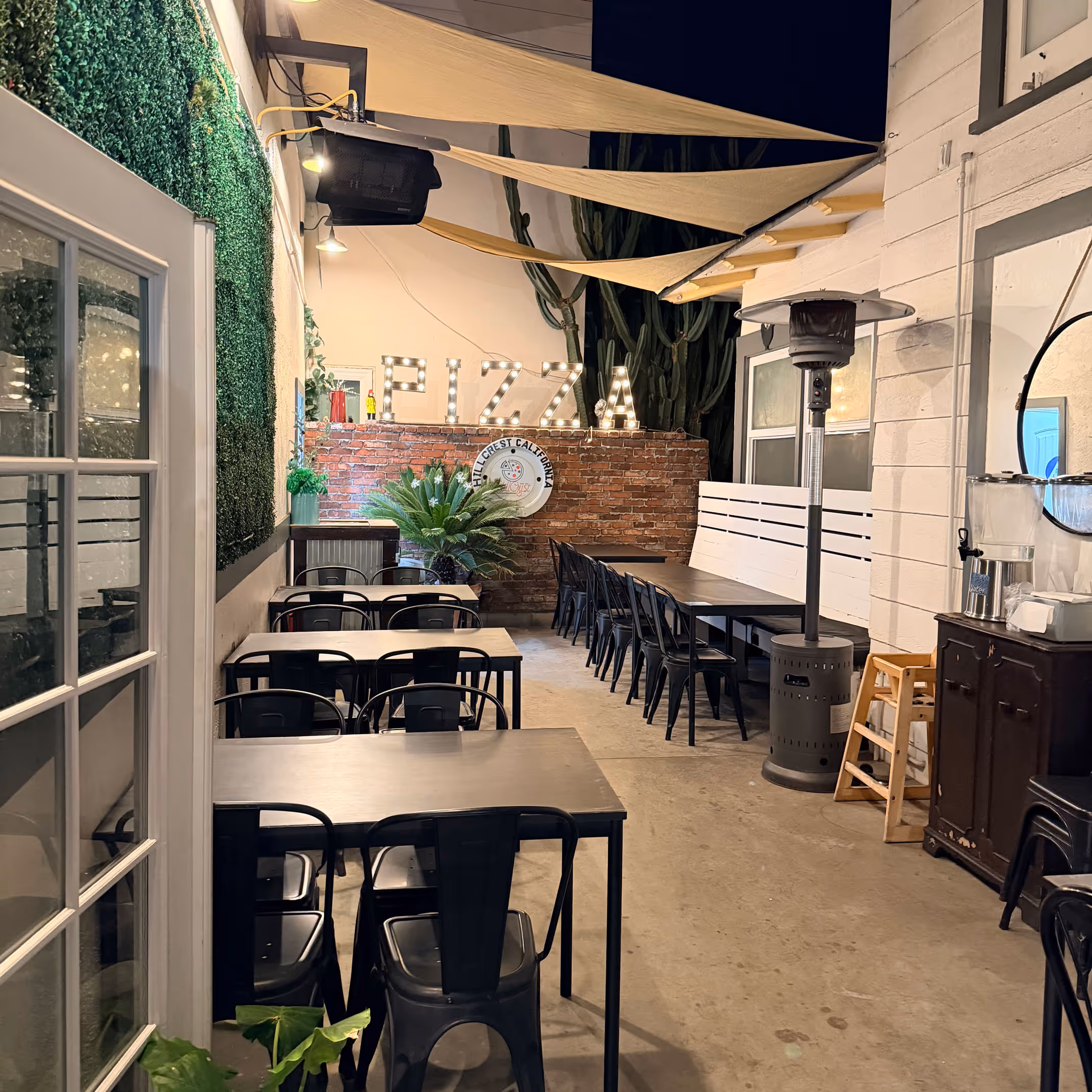 Outdoor pizza patio with string lights spelling "PIZZA," black tables and chairs, a brick wall, and lush greenery. Cozy, inviting atmosphere.