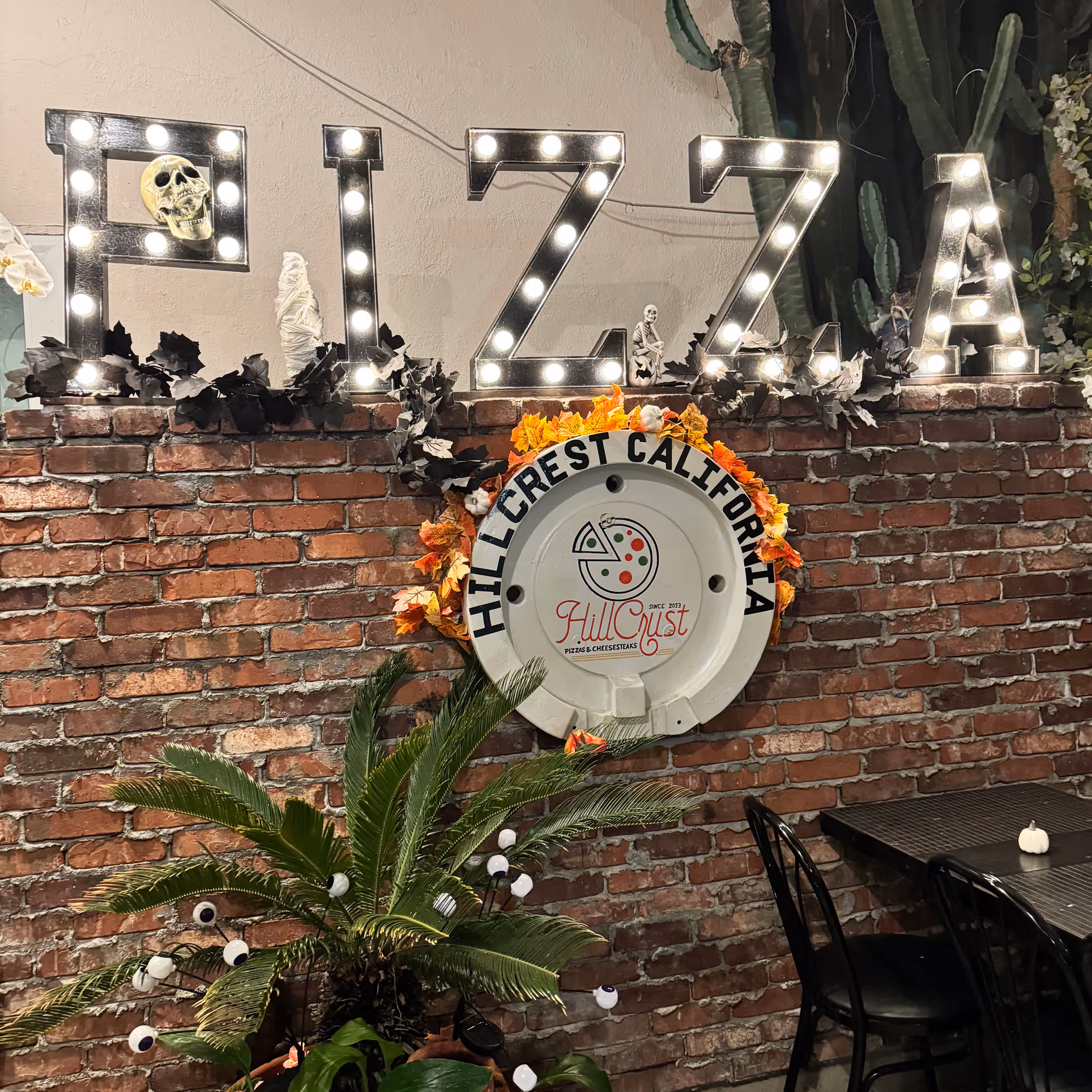Illuminated "PIZZA" sign on a brick wall with Halloween decor, featuring skulls and dark leaves. Below, a "Hillcrest California" logo surrounded by fall leaves, next to a plant and empty chairs.