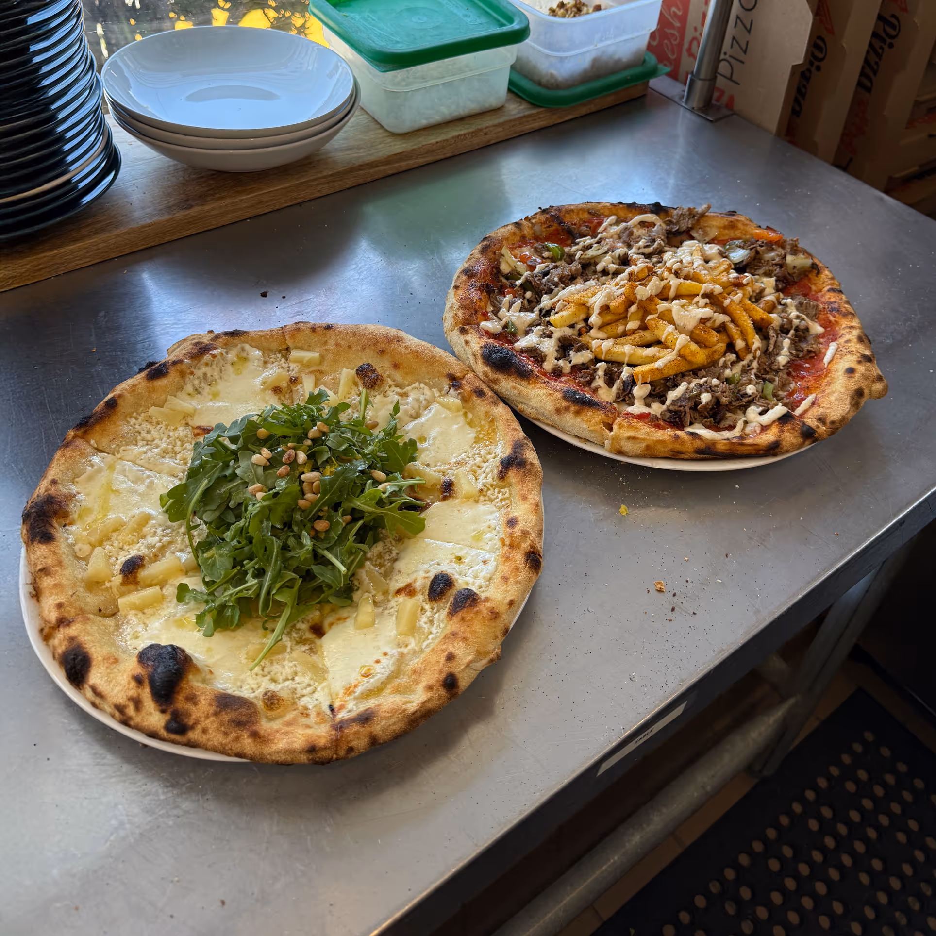 Two pizzas on a metal counter: one with arugula, pine nuts, and cheese; the other with beef, fries, and sauce, creating a rustic, appetizing scene.