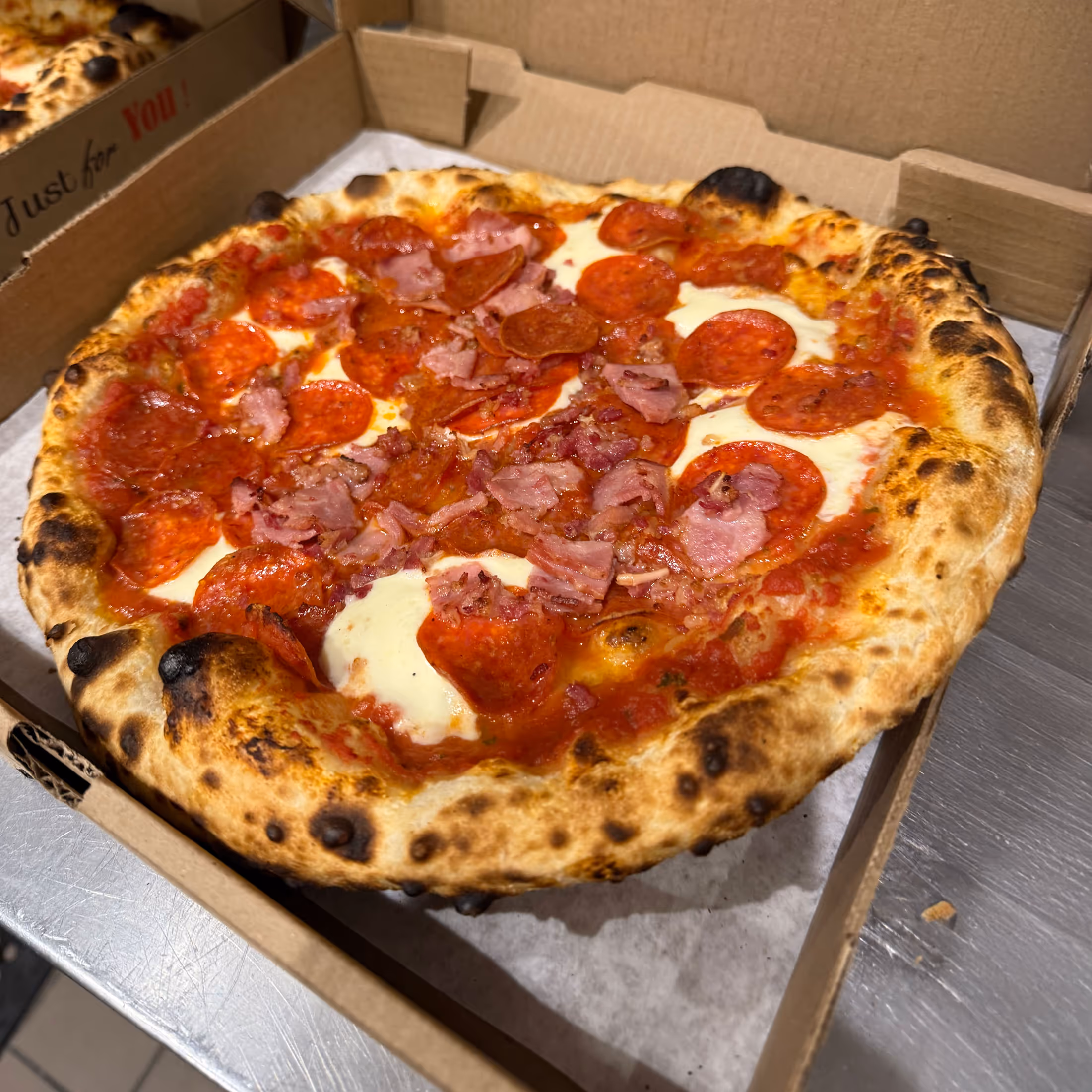 Wood-fired pizza with a golden crust, topped with pepperoni, bacon, and melted cheese, rests in an open cardboard box on a countertop.