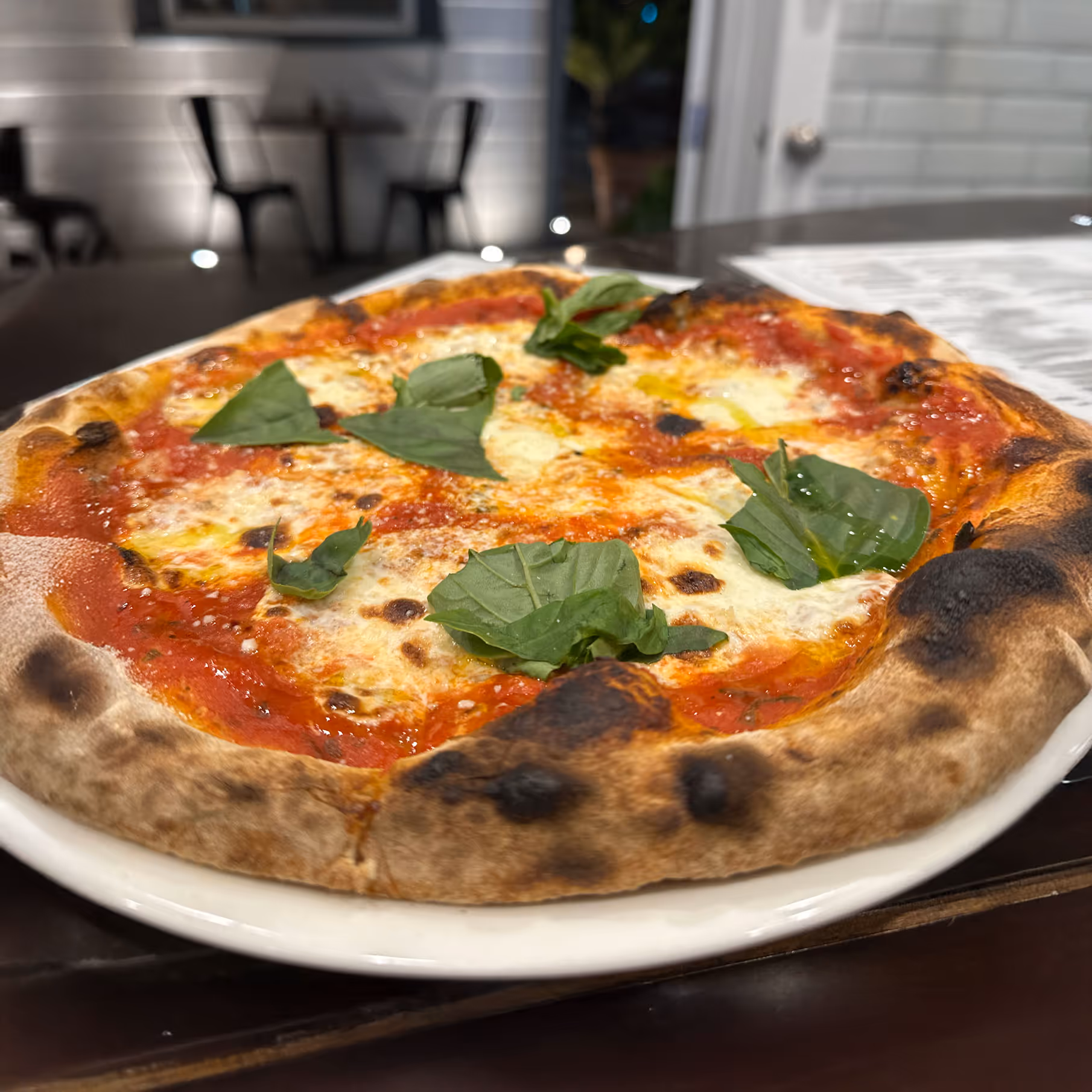 Close-up of a Neapolitan pizza with vibrant red sauce, melted cheese, and fresh basil leaves, served on a white plate in a cozy pizzeria setting.