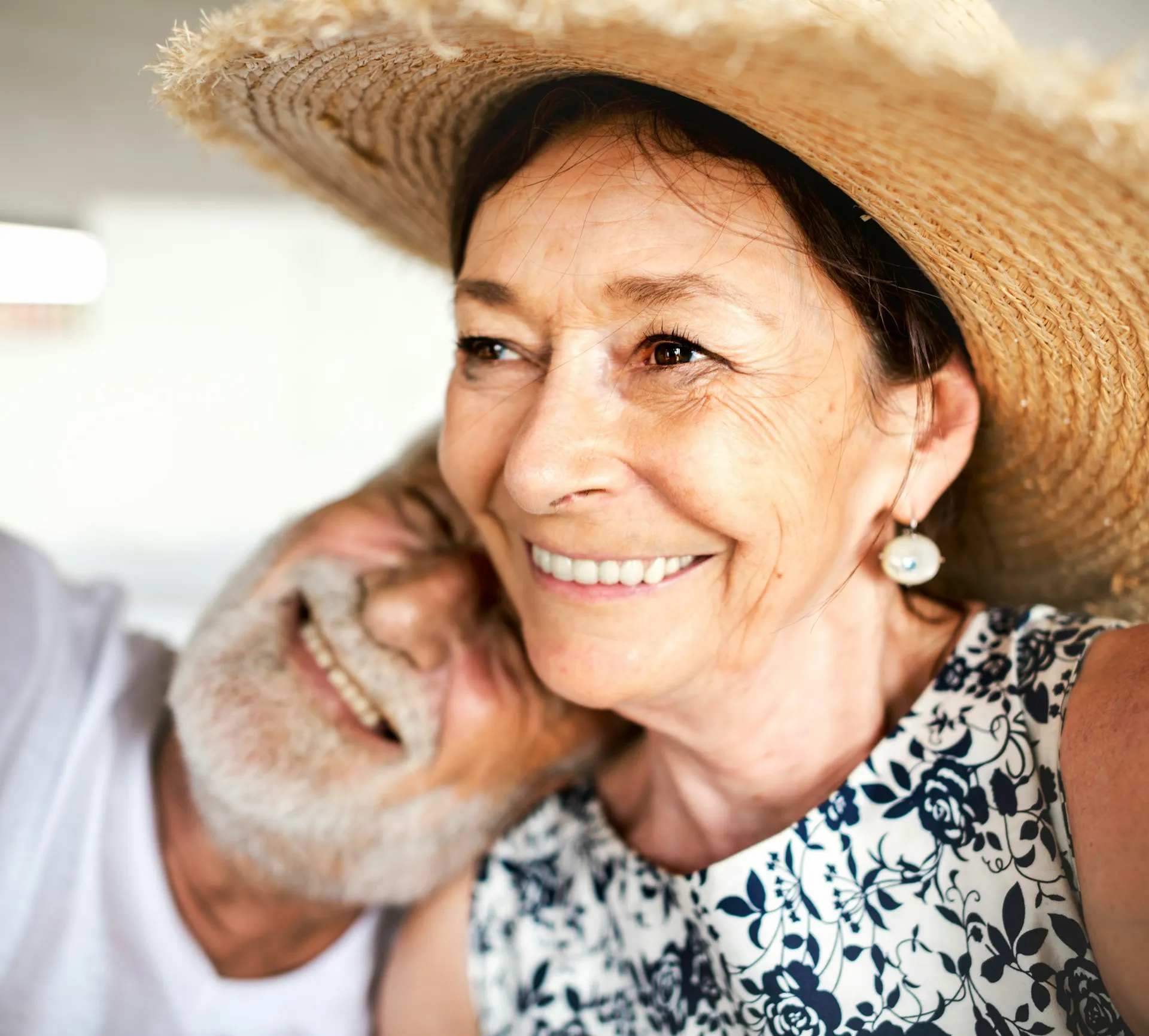 Close-up of a smiling older woman wearing a straw hat and floral dress, with a bearded older man resting his head on her shoulder.