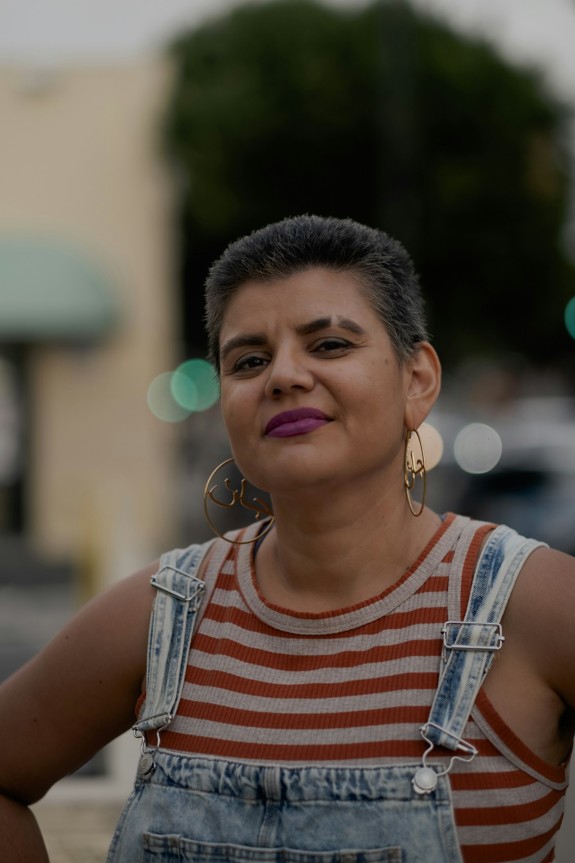 Confident person with short dark hair wearing large hoop earrings, a striped sleeveless top, and denim overalls.