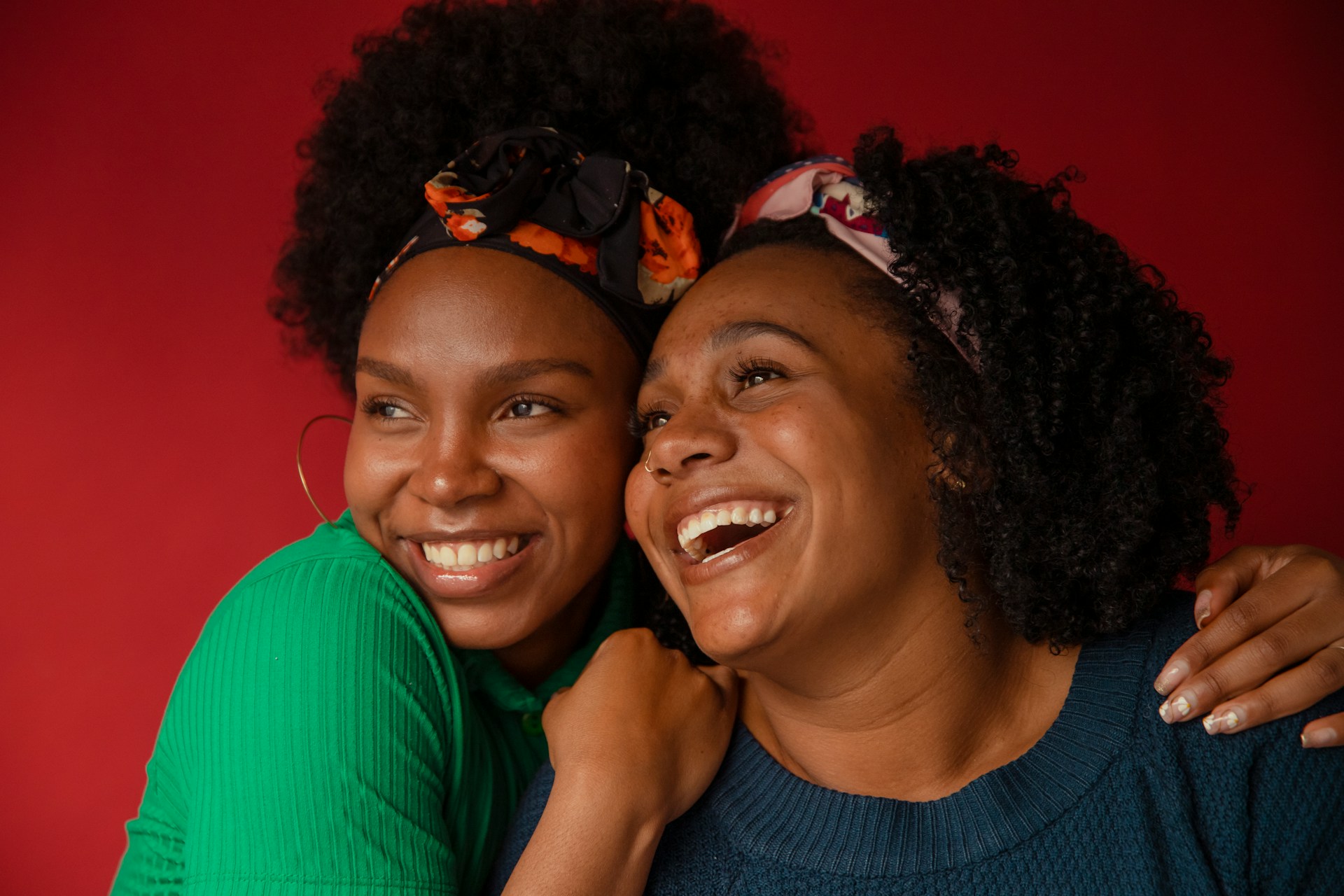 Two women smiling and hugging against a red background, both wearing colorful headbands and sweaters.