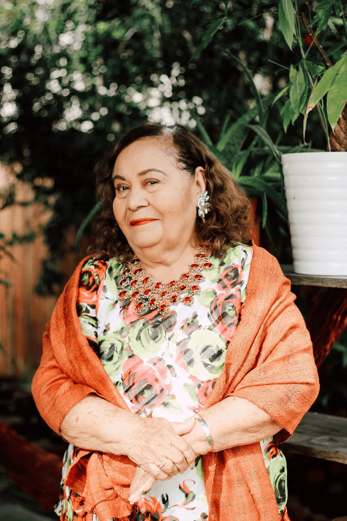 Elderly woman wearing a floral dress, orange shawl, red statement necklace, and large earrings standing outdoors with greenery in the background.