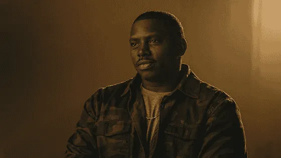 Portrait of a thoughtful young man wearing a dark jacket and chain necklace, illuminated by warm light against a neutral background.