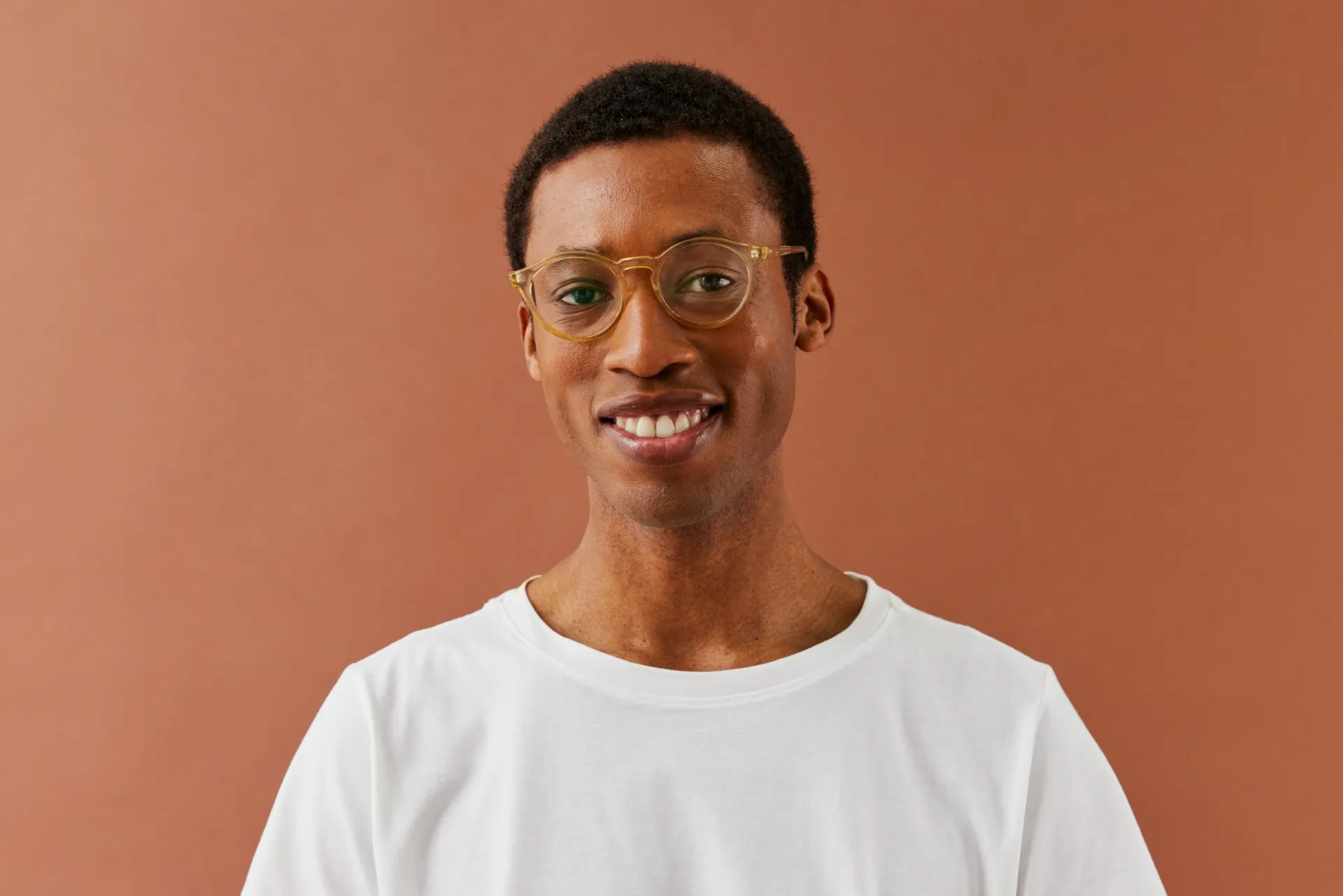 Smiling young man with short curly hair and clear round glasses wearing a white shirt against a brown background.