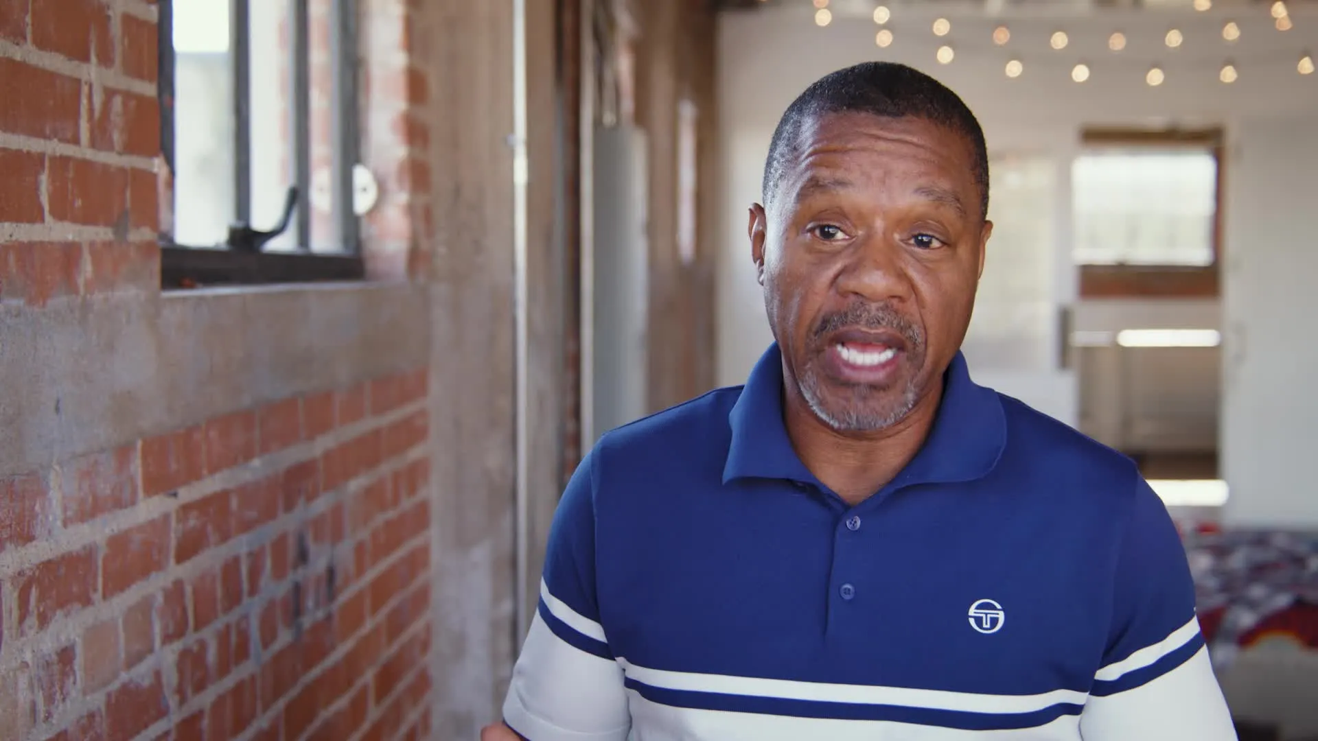 Middle-aged man in a blue and white striped polo shirt speaking indoors in front of a brick wall with string lights in the background.