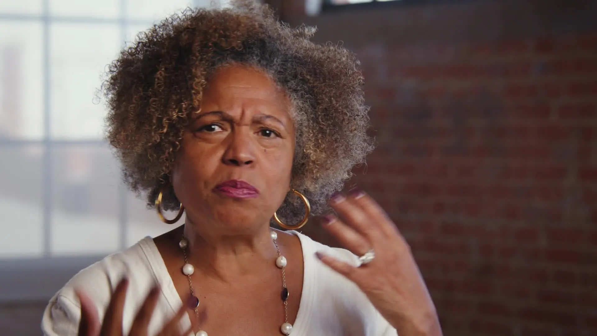 Woman with curly hair and gold hoop earrings speaking expressively indoors.