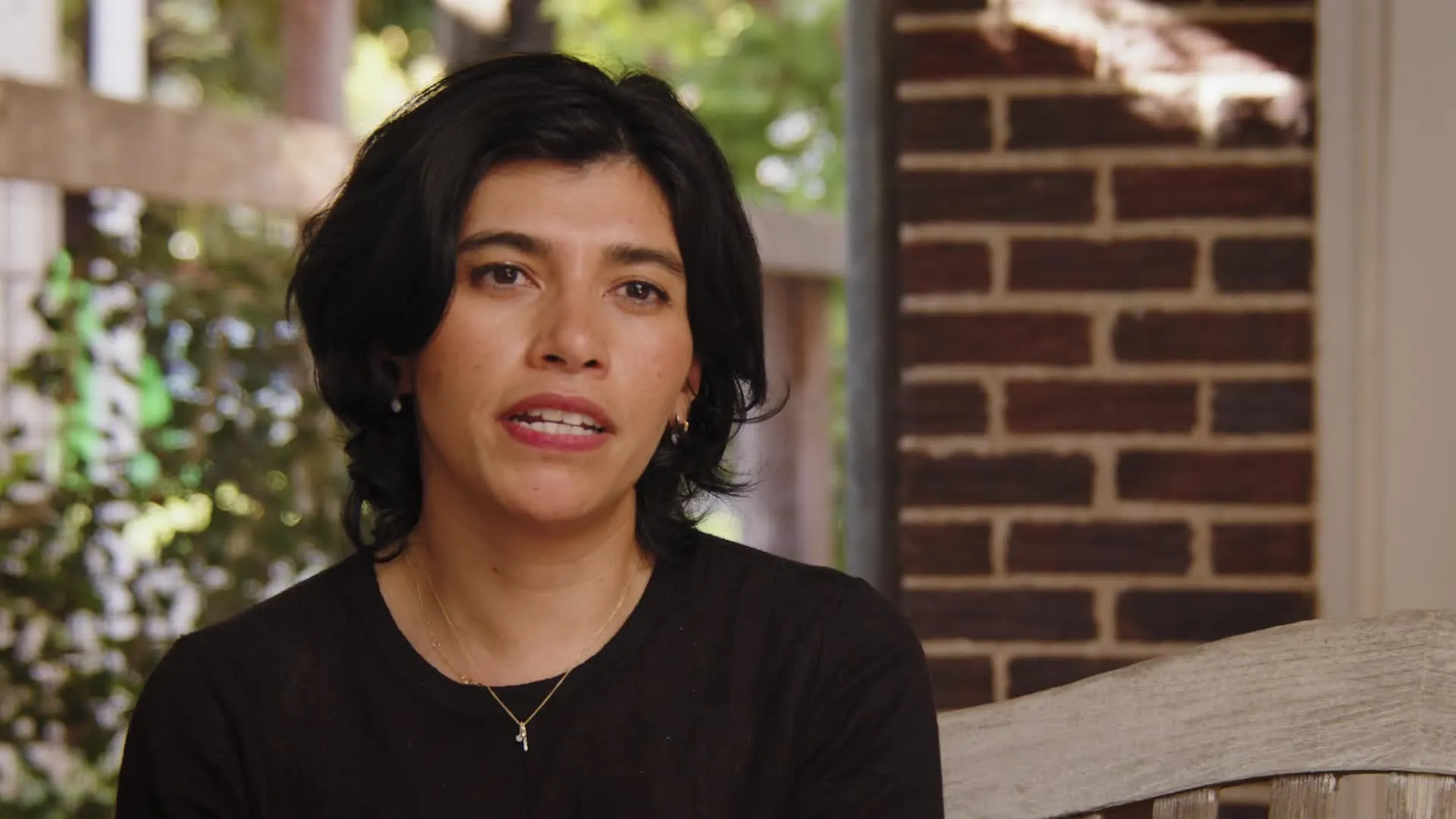 Woman with short black hair wearing a black top, sitting indoors near a brick wall with greenery visible outside.