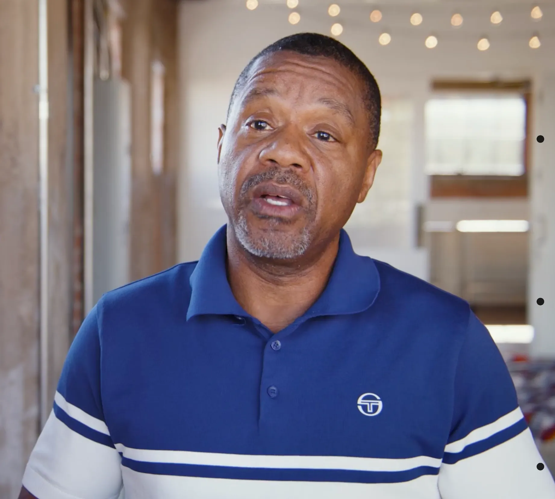 Middle-aged man in a blue and white striped polo shirt speaking indoors in front of a brick wall with string lights in the background.