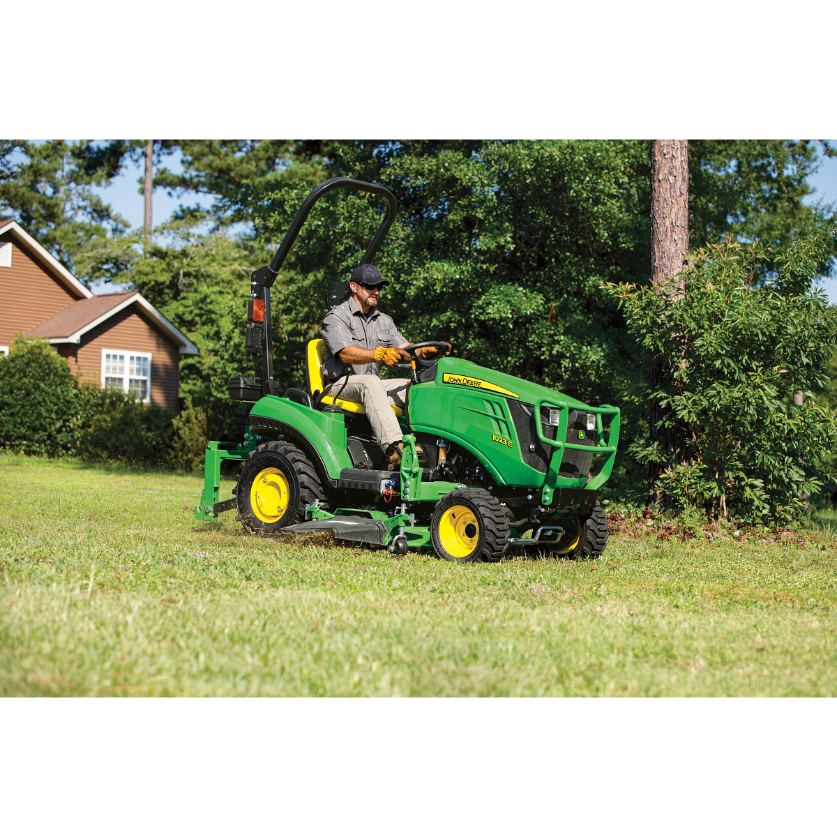 Person operating a green compact tractor with a mower attachment cutting grass across a yard near a house, with trees and shrubs in the background