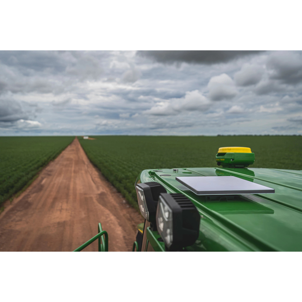 JDLink Boost Mini Terminal mounted on the roof of a John Deere machine, viewed from above with a dirt road, crop fields, and cloudy sky stretching into the distance.