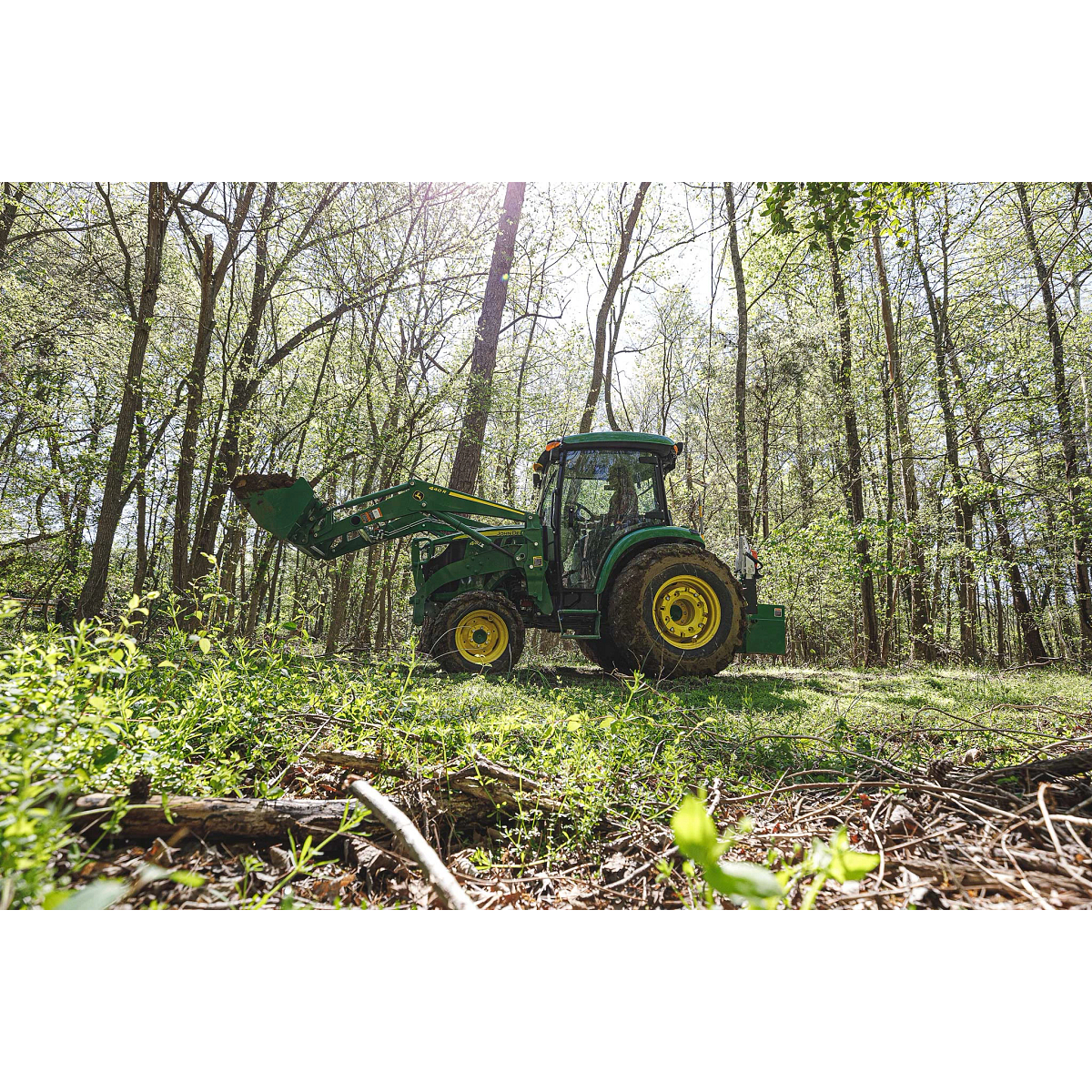Side left view of a 4044R Compact Utility Tractor driving through a forest with loader extended, carrying a full load of dirt.
