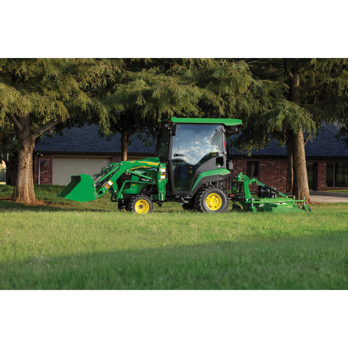 A green tractor with an enclosed cab operates in a grassy yard, using a rear mower attachment to cut grass with a building and large trees in the background