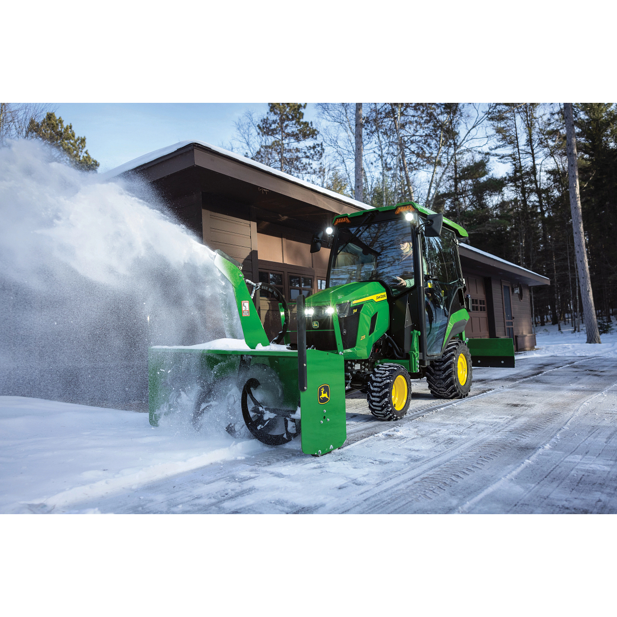 A green tractor equipped with a front-mounted snow blower clears snow from a driveway in front of a garage, blowing a wide spray of snow to the side among tall trees
